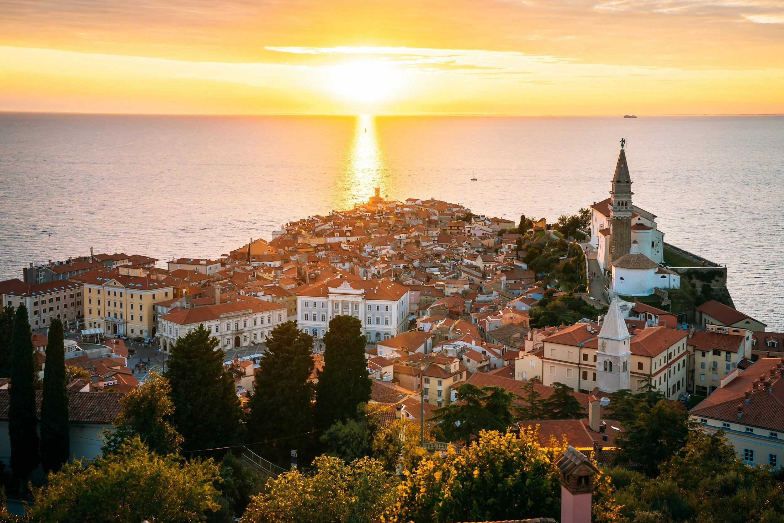 Sunset over a coastal town with red-roofed buildings, a church with a tall steeple, and the sea in the background.