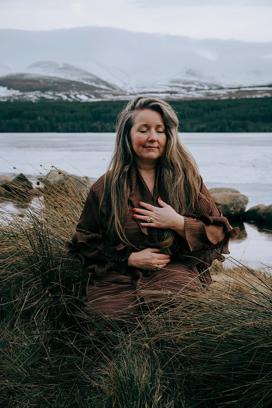 Rev. Bunny Love-Schock sitting in the tall grass by a lake with her eyes close and her hand over her heart