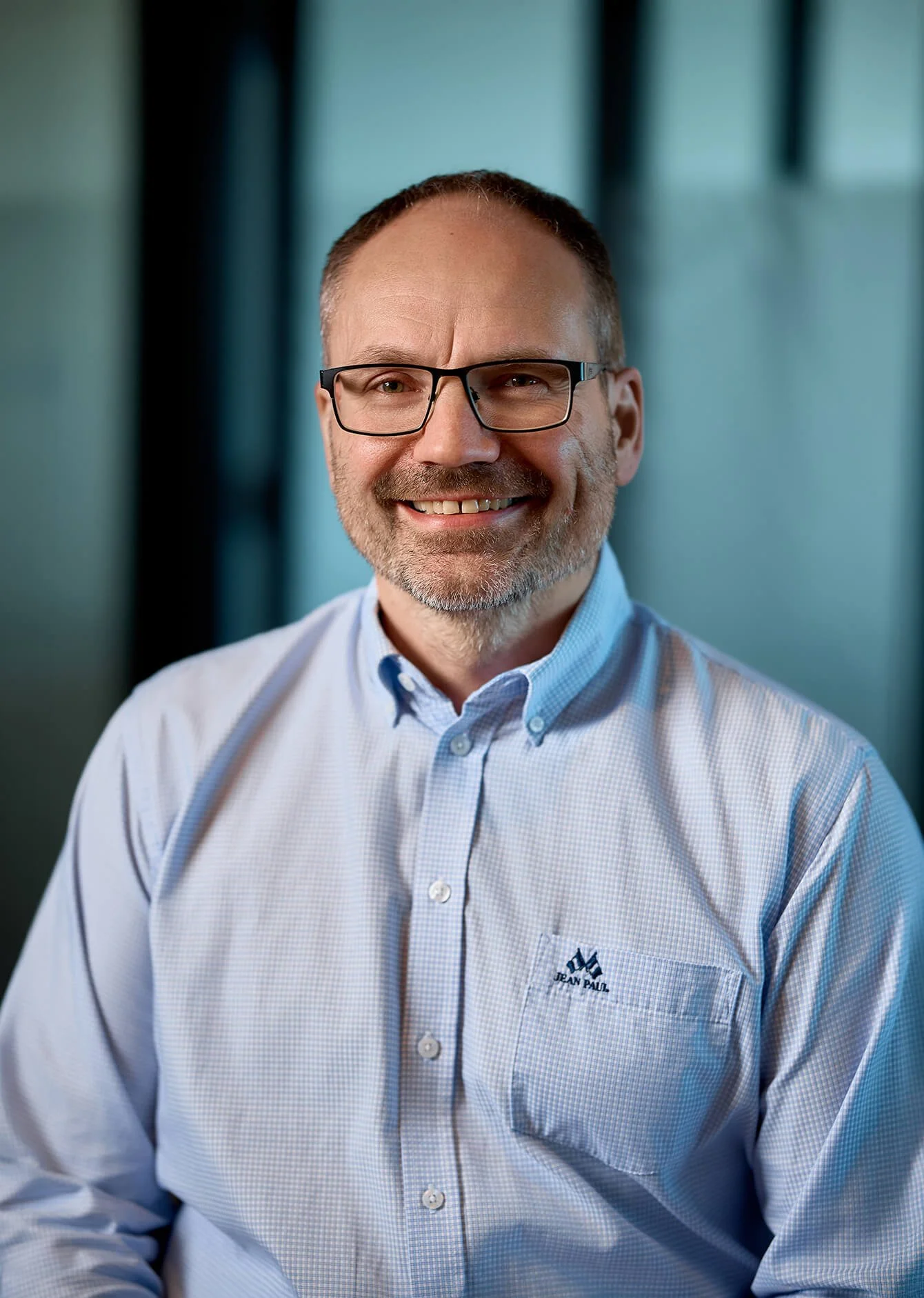 A smiling man with glasses, wearing a light blue checkered dress shirt with a Jean Paul Gaultier logo on the chest pocket, sitting against a blurred background with blue and green hues.