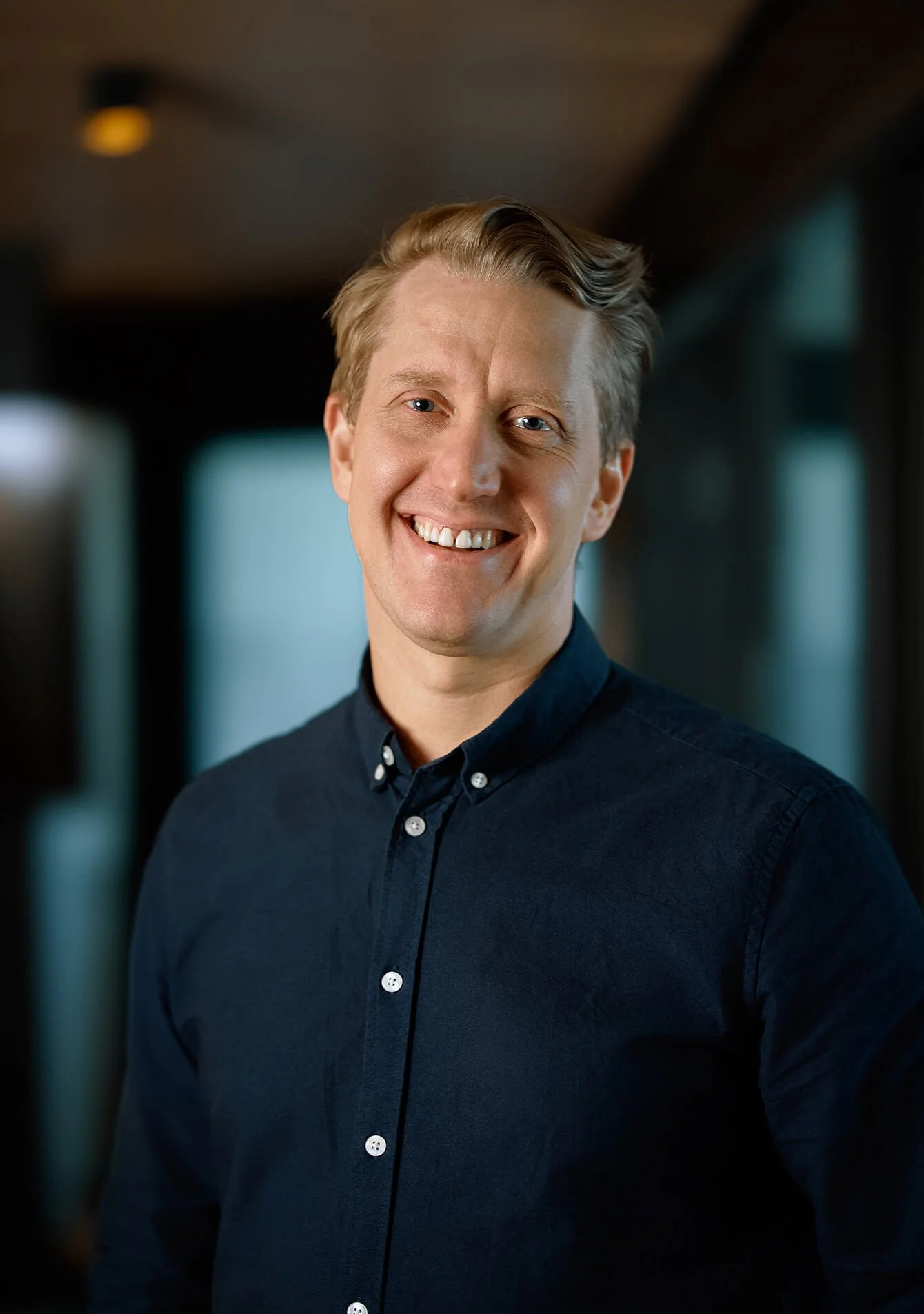 A smiling young man with light blond hair, blue eyes, and fair skin, wearing a dark blue button-up shirt, standing indoors with a blurred background.