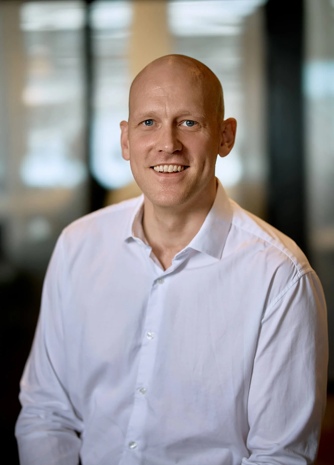 Portrait of a smiling man with a shaved head, wearing a white dress shirt, in a blurred indoor background.