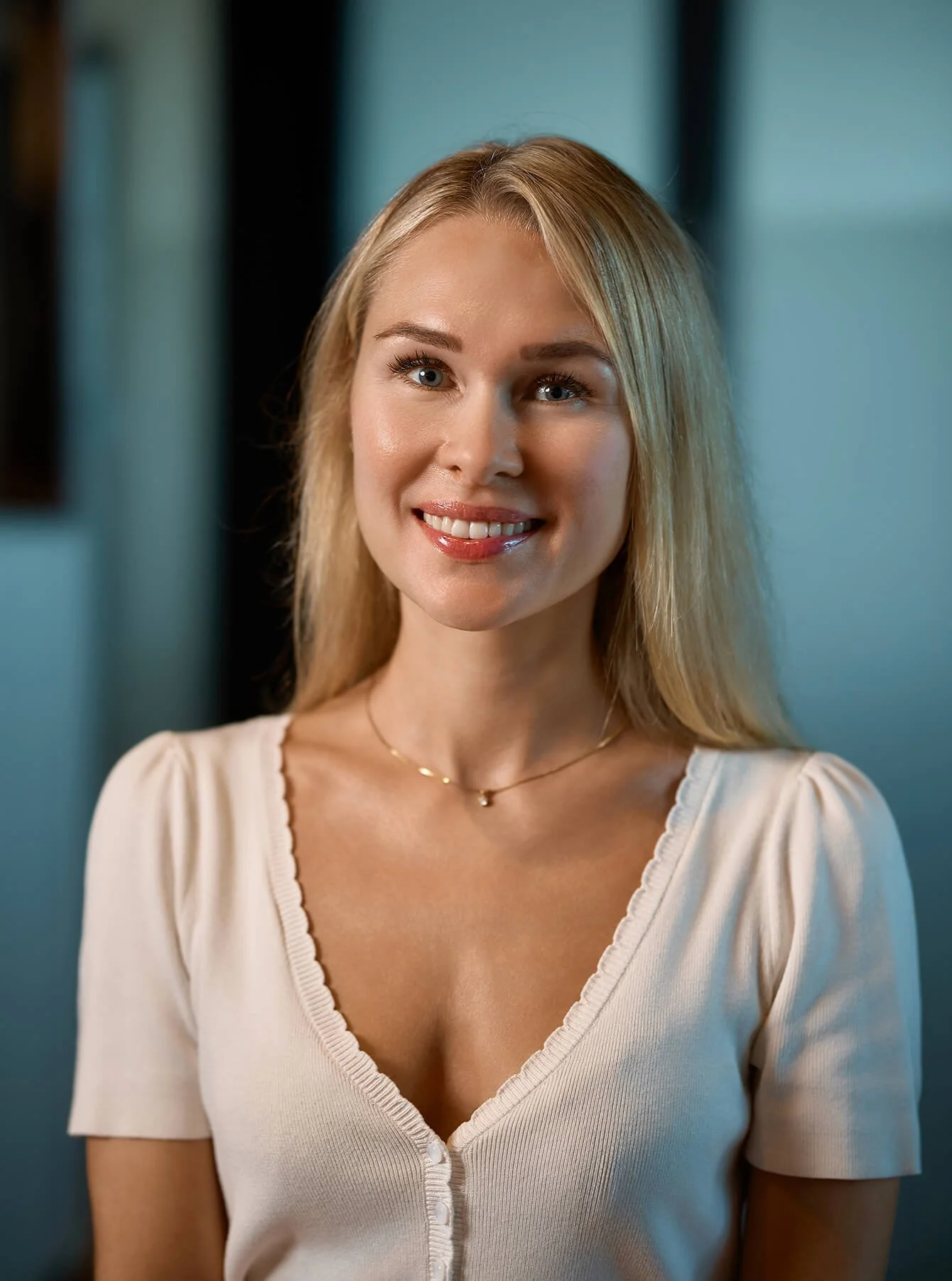 Close-up of a young woman with blonde hair, wearing a cream-colored top with a v-neck and buttons, smiling against a-colored background.