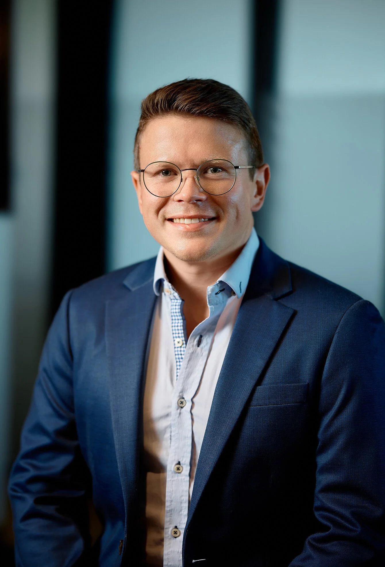 Portrait of a young man with short brown hair, wearing round glasses, a white patterned shirt, and a navy blazer, smiling at the camera in a professional setting.