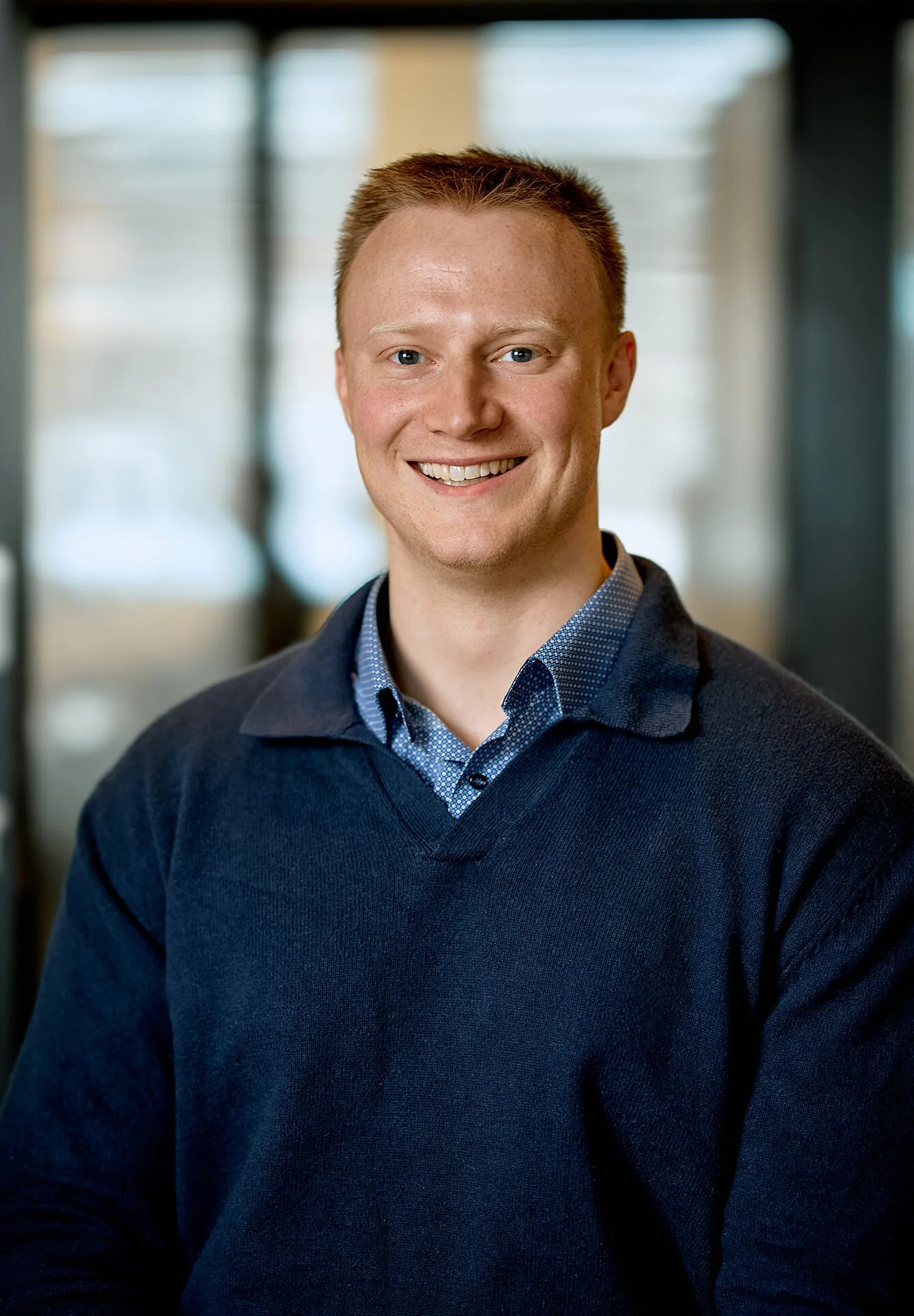Portrait of a smiling young man with short reddish hair, wearing a blue collared shirt under a dark sweater, in an indoor setting with blurred background.