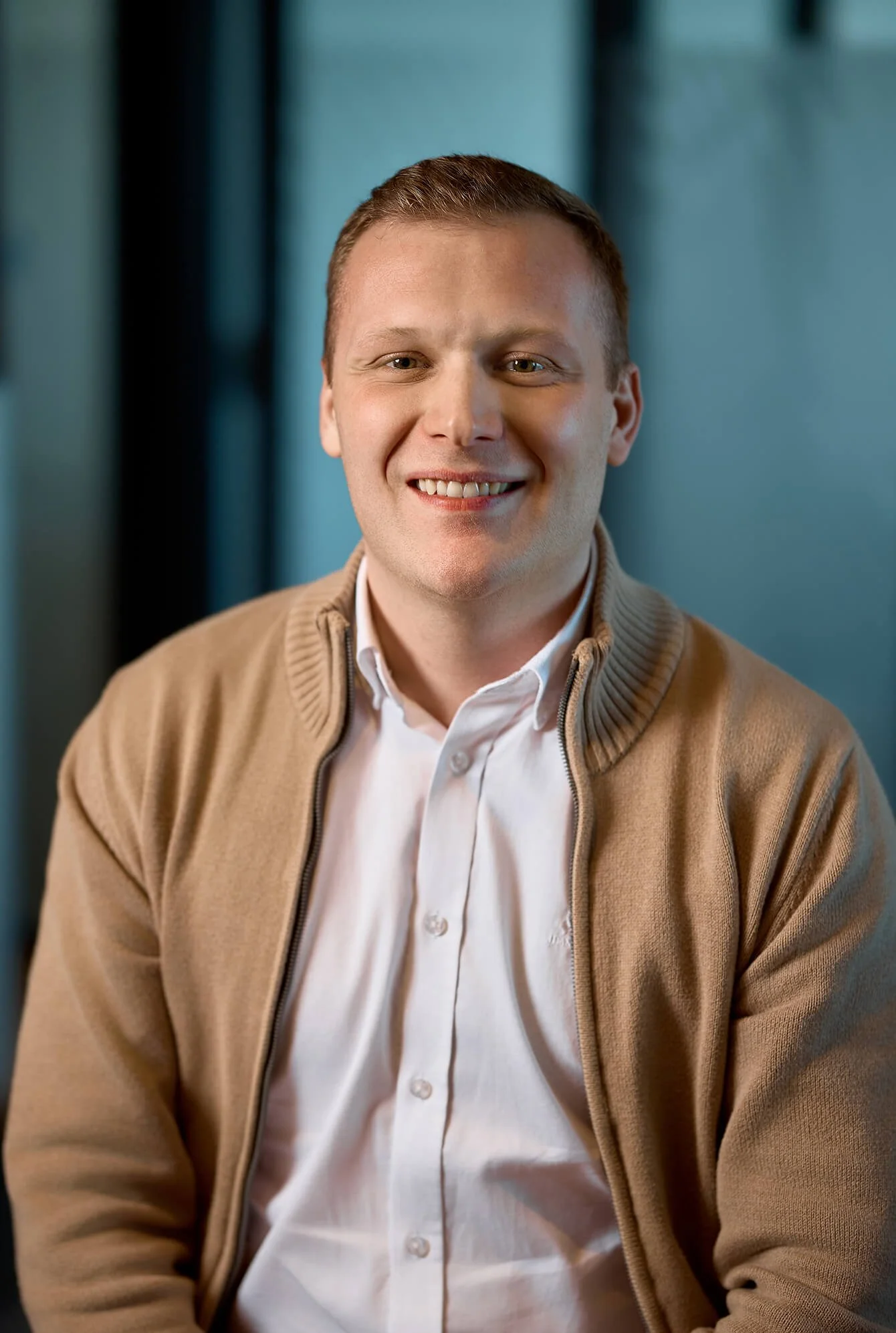 A smiling man wearing a beige zip-up sweater over a white button-up shirt, sitting indoors with a blurred blue background.