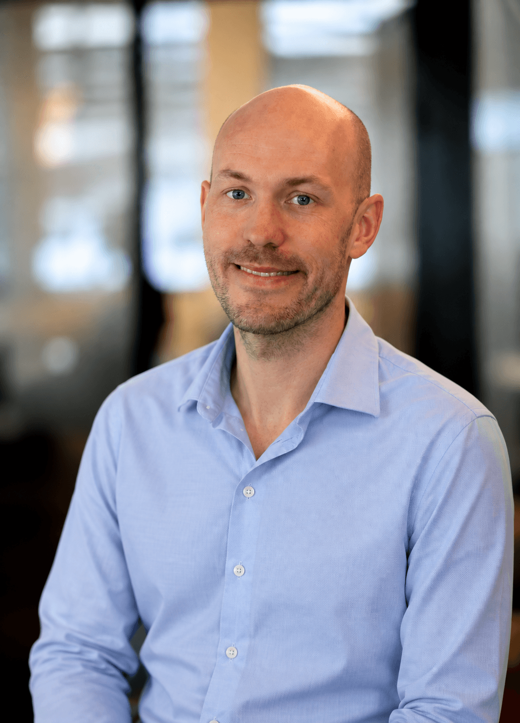 Headshot of a bald man with a trimmed beard and mustache, wearing a light blue button-up shirt, smiling, in an indoor setting with blurred background.