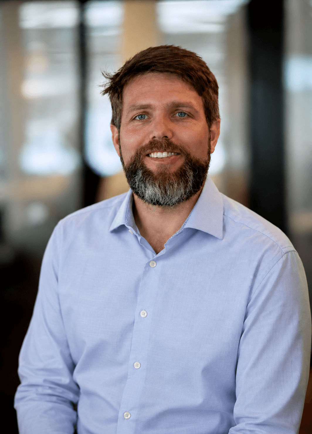 Headshot of a man with brown hair and a beard, wearing a light blue button-down shirt, smiling with a blurred indoor background.