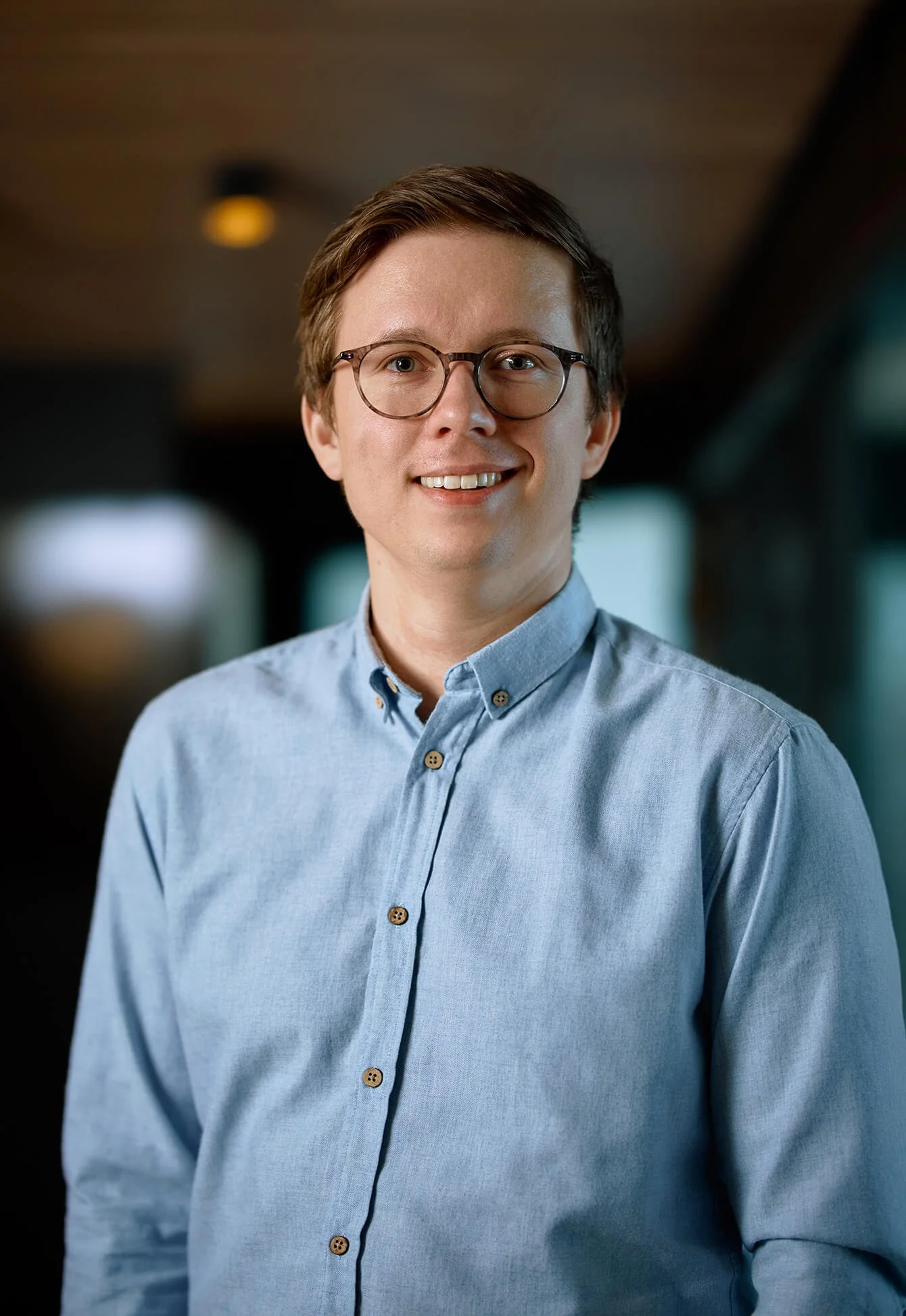 A young man with glasses and light brown hair, wearing a light blue button-up shirt, smiling indoors with blurred background.