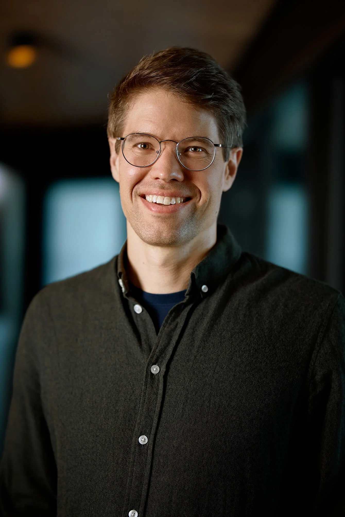Portrait of a smiling man with short brown hair, wearing glasses and a dark button-up sweater, standing indoors.