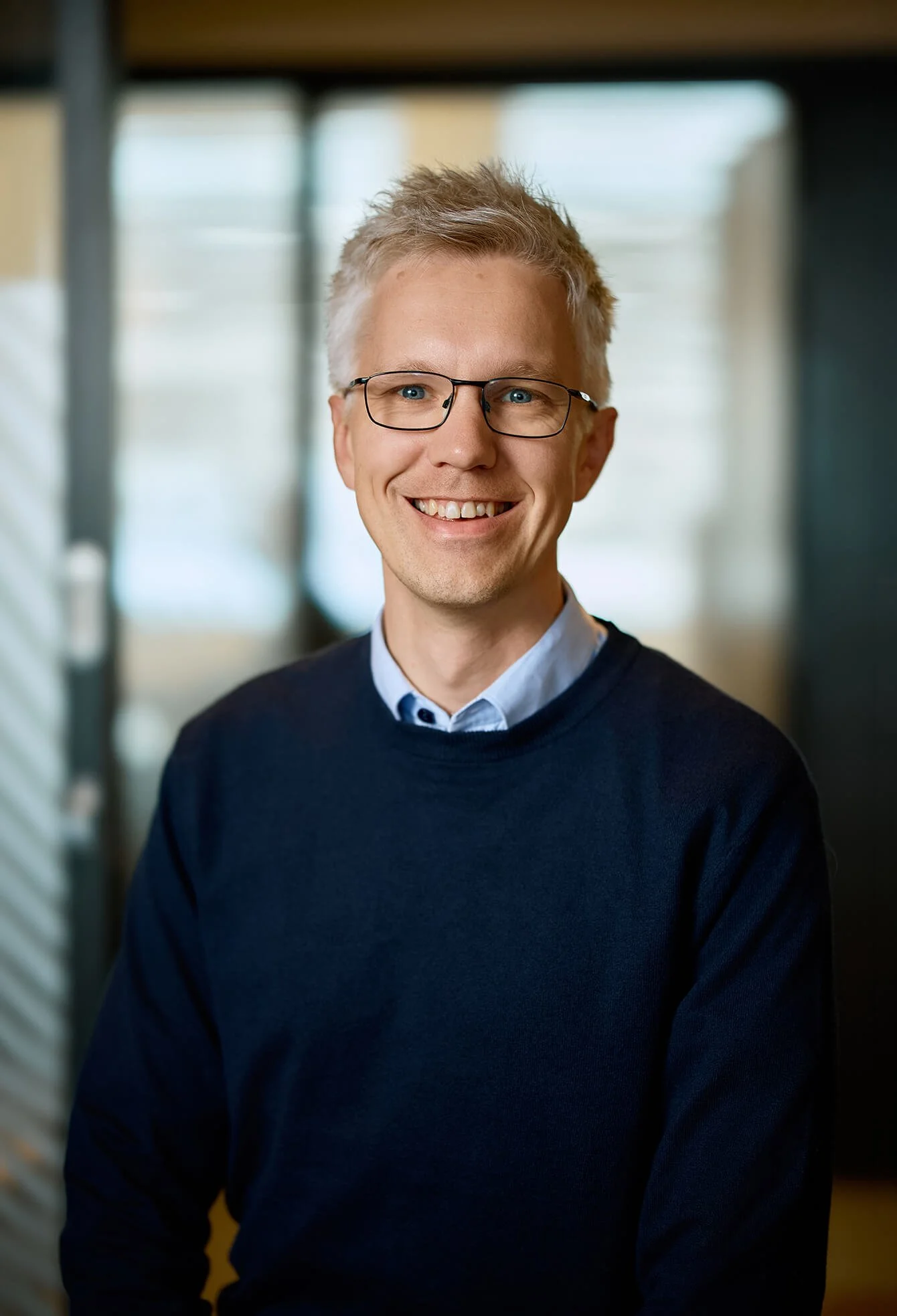 A smiling man with glasses and blond hair wearing a blue sweater, standing indoors in front of windows.