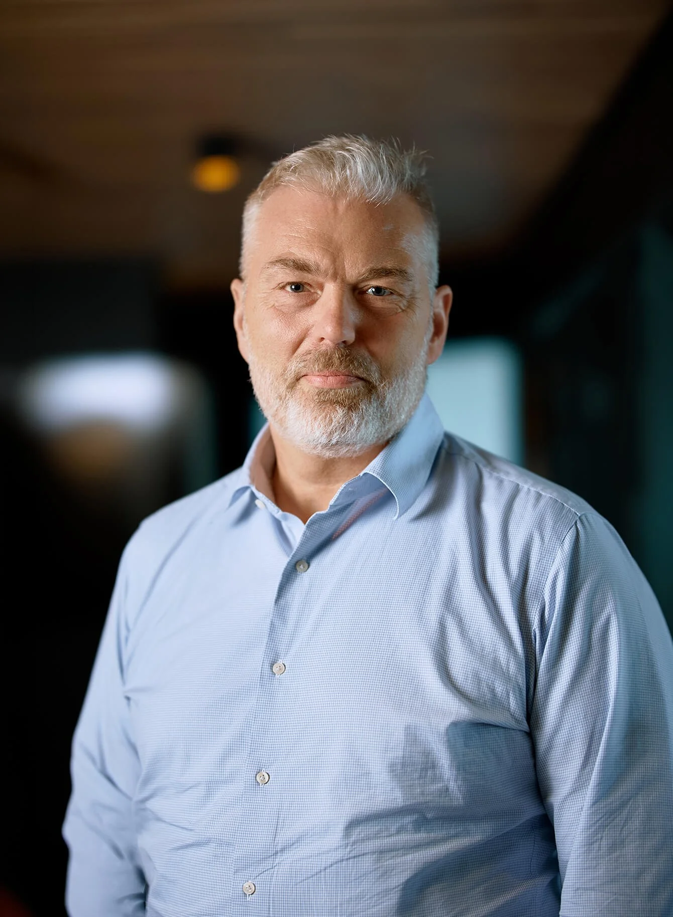 A middle-aged man with gray hair and a beard, wearing a light blue dress shirt, standing indoors with blurred background.