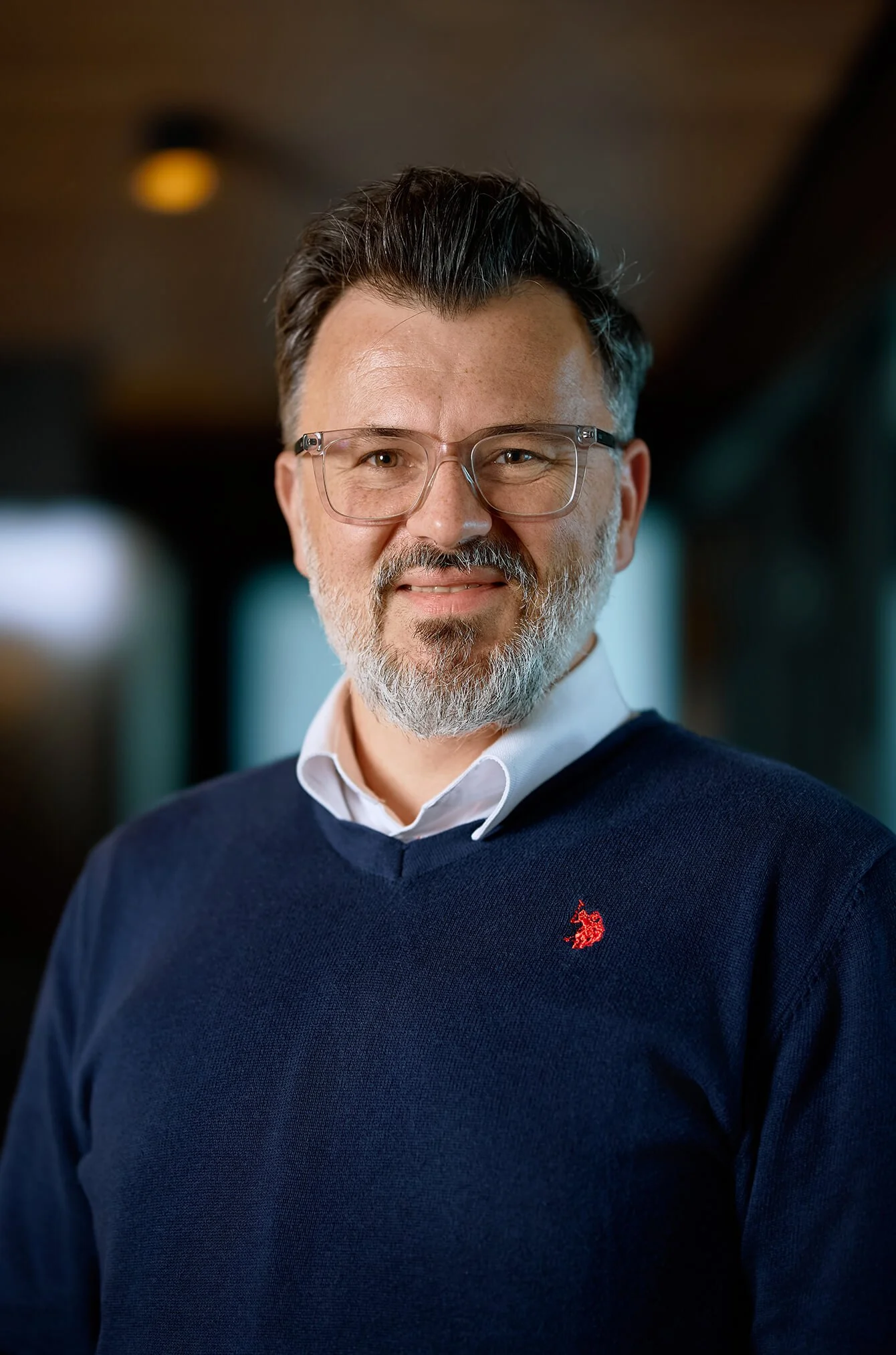 A man with glasses and gray hair, wearing a navy blue sweater with a red logo, smiling indoors with blurred background.