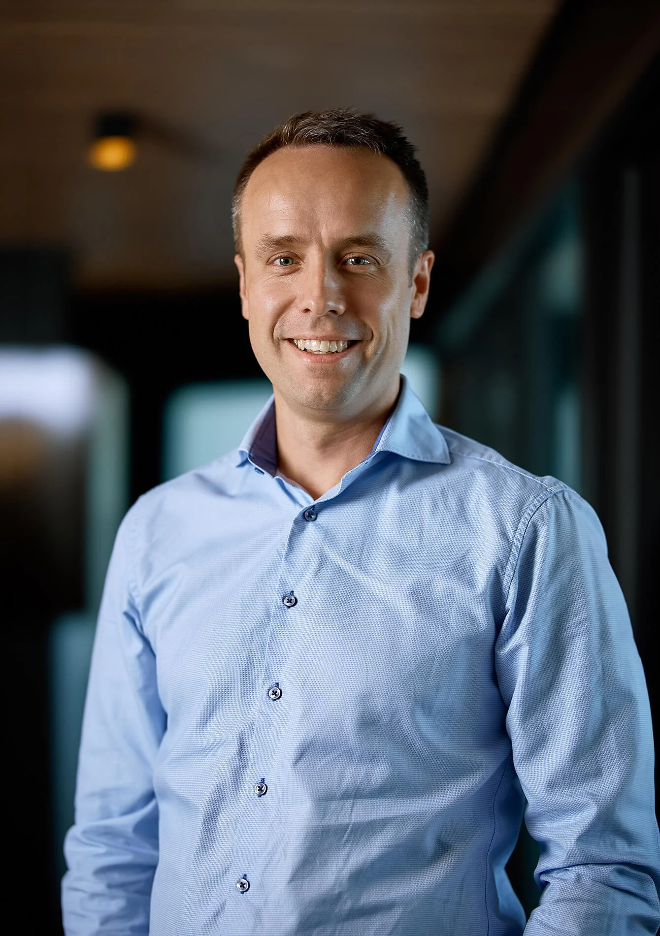 A man smiling, wearing a light blue button-up shirt, with a blurred background that appears to be the inside of a vehicle.