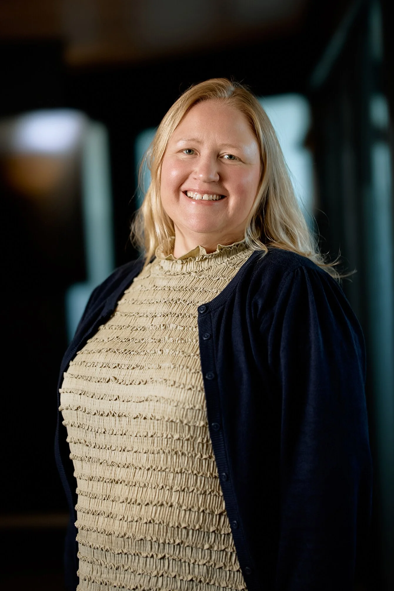 A smiling woman with blonde hair, wearing a beige ruffled top and a navy cardigan, standing indoors with a blurred background.