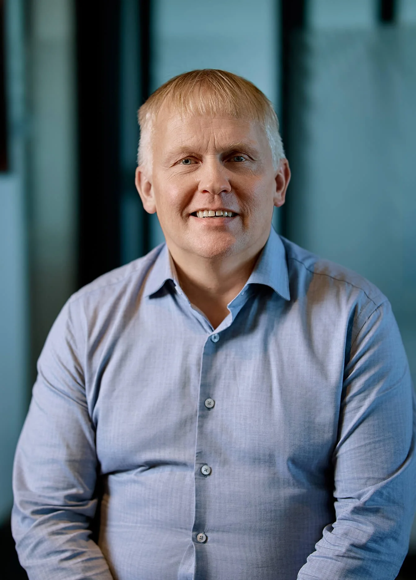 A portrait of a middle-aged man with short blond hair, wearing a light blue button-up shirt, smiling, sitting in front of a blurred background with shades of blue and black.
