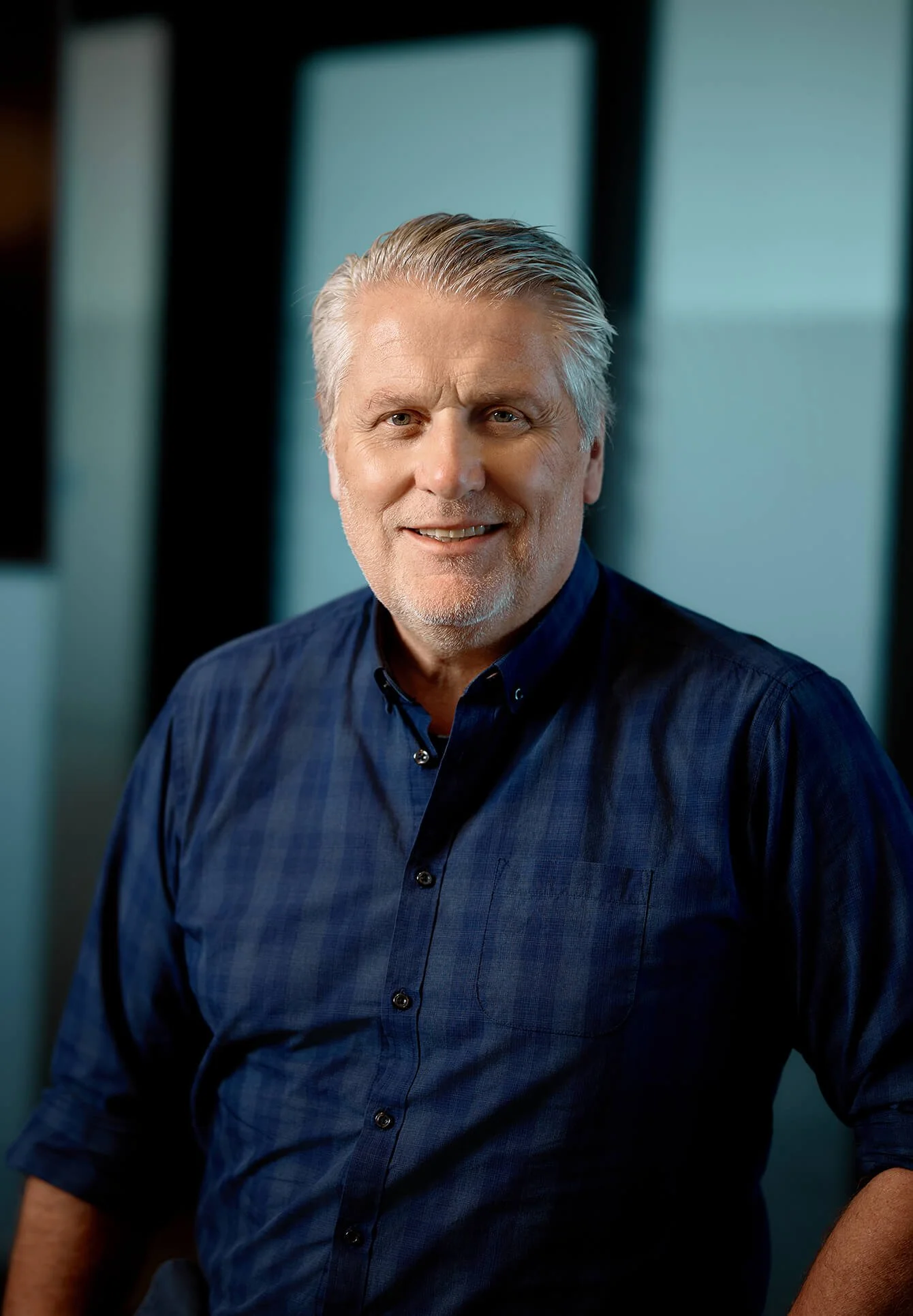 A middle-aged man with gray hair and blue eyes smiling, wearing a dark blue button-up shirt, seated in front of a blurred background with light blue panels.