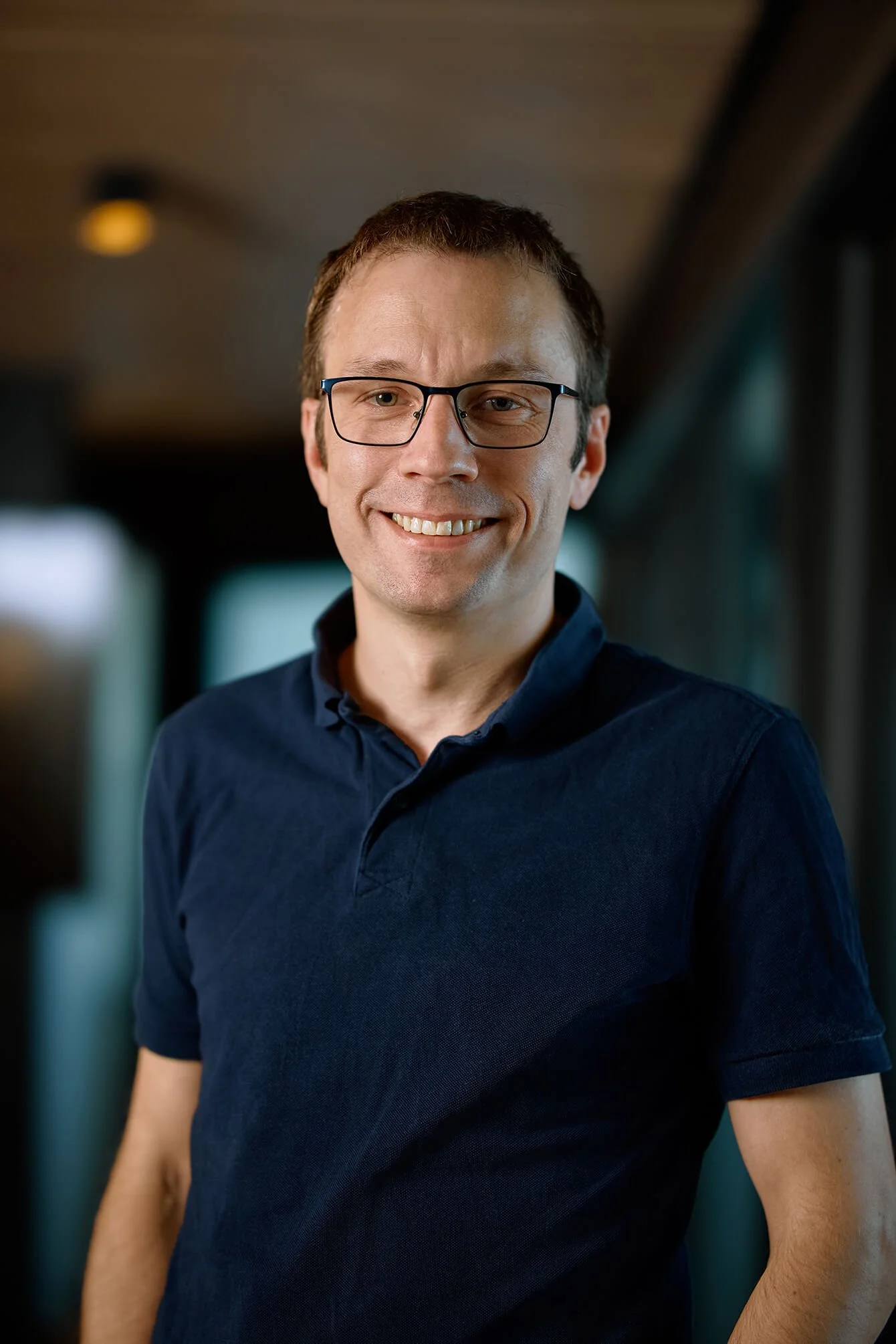 A smiling man with glasses and short brown hair wearing a navy blue polo shirt.