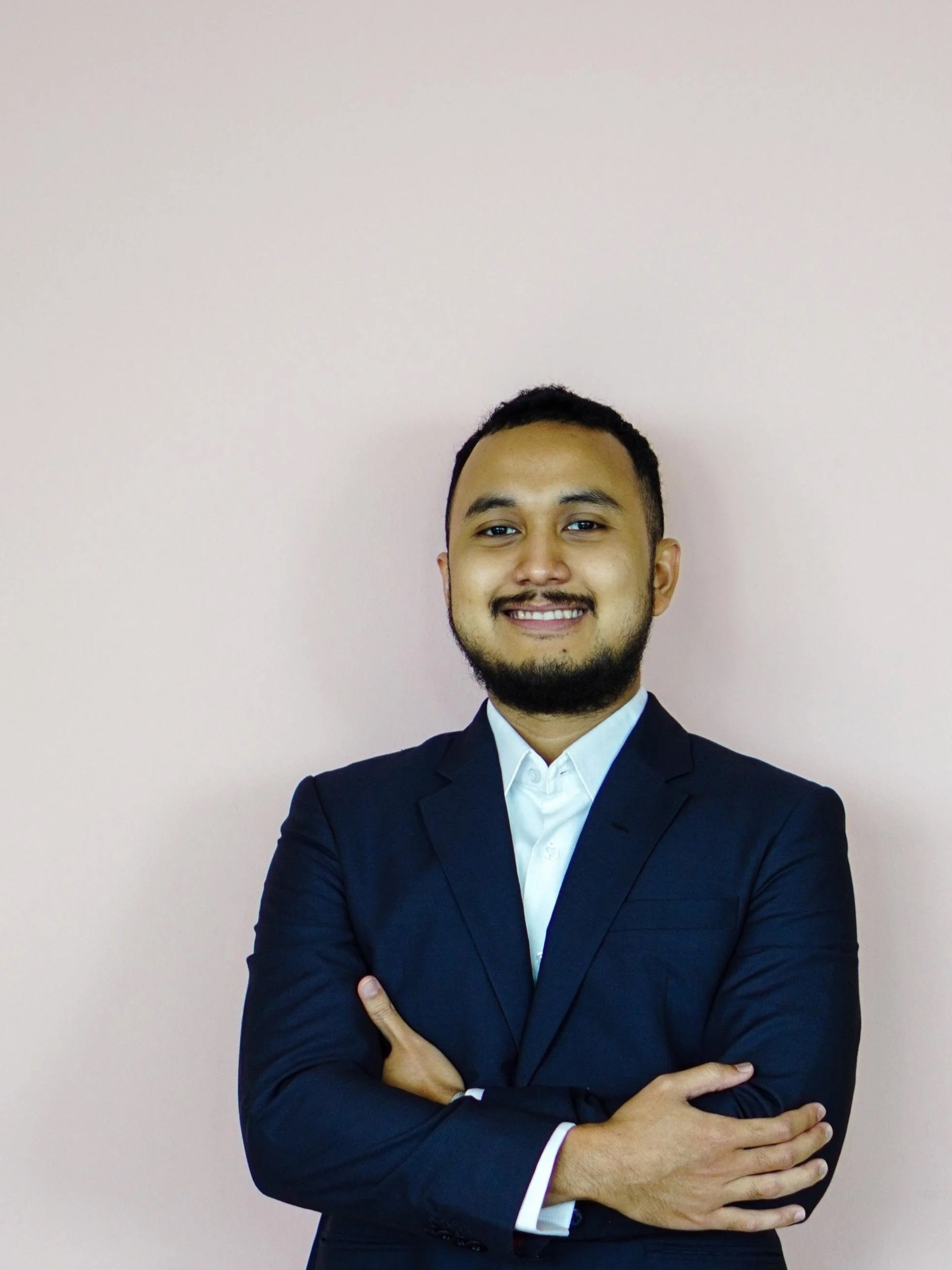 An Indonesian man smiling in front of a pink wall.
