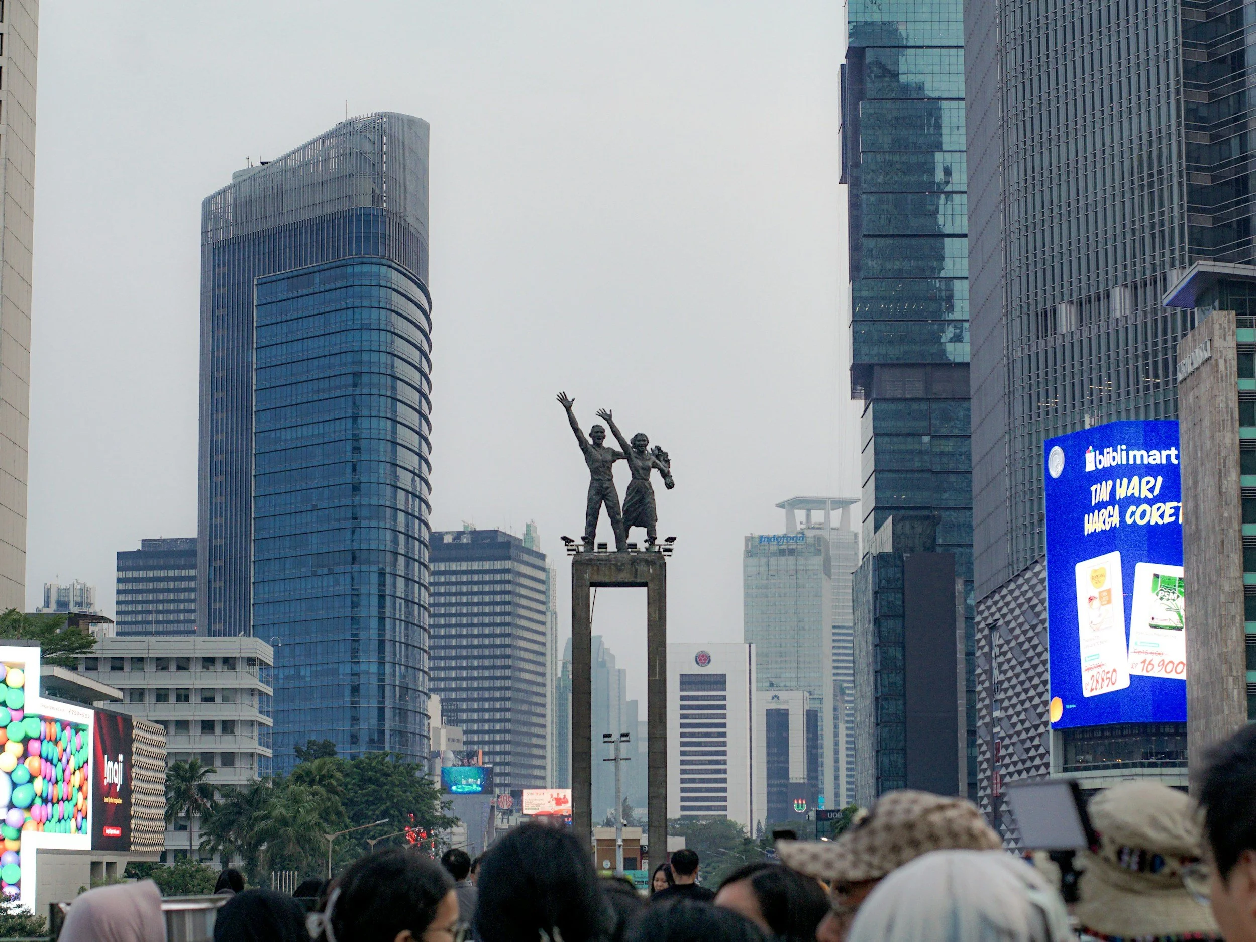 Crowd of people gathered in the Welcome Statue in Jakarta's Bundaran HI with tall modern skyscrapers in the background. There is a statue of two figures holding hands in the center, and billboards with advertisements around the area.