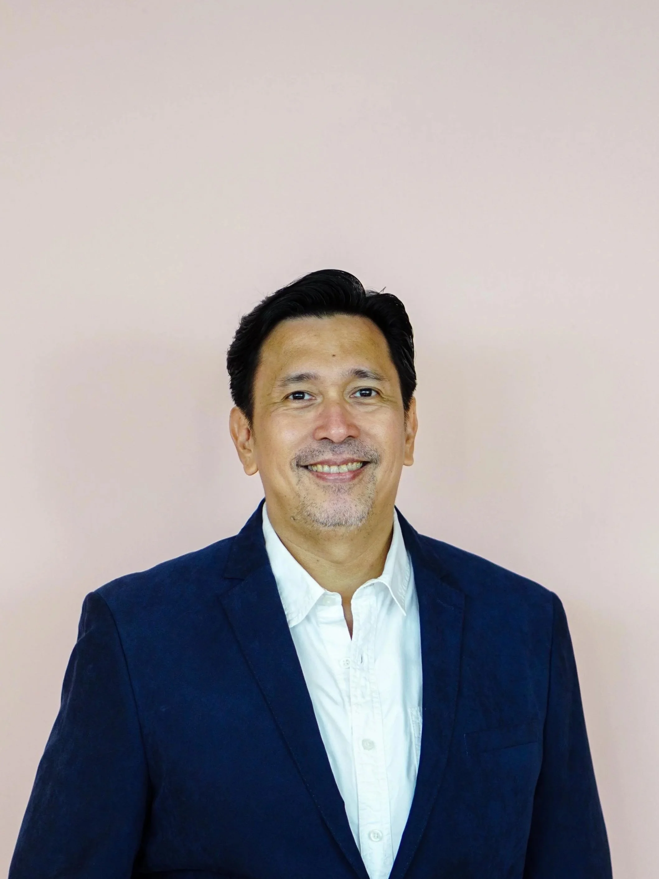 An Indonesian man wearing a navy blazer over white shirt is standing in front of a pink wall.