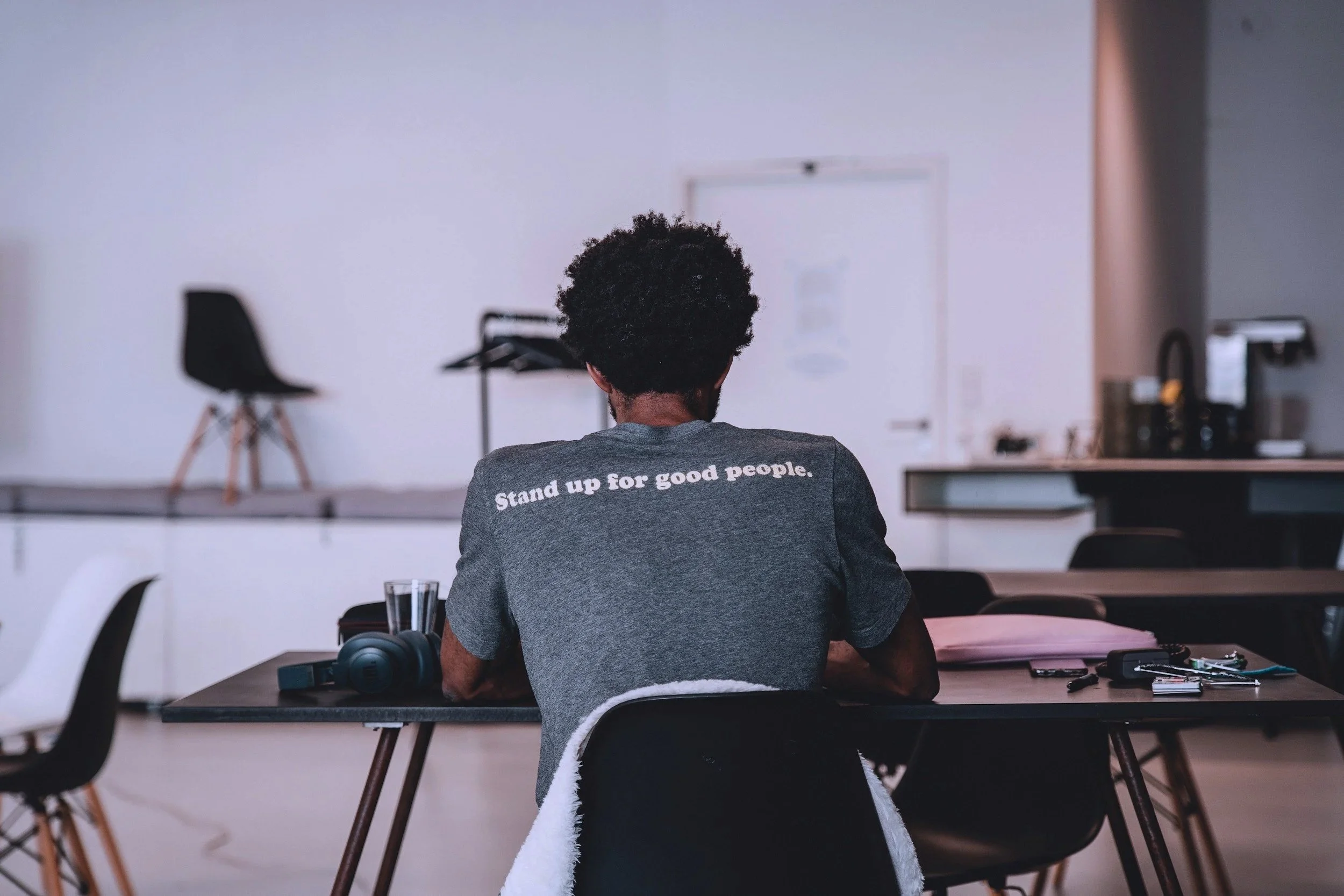 Back of a person with curly hair sitting at a desk in a meeting or classroom, with a white wall and a door in the background.