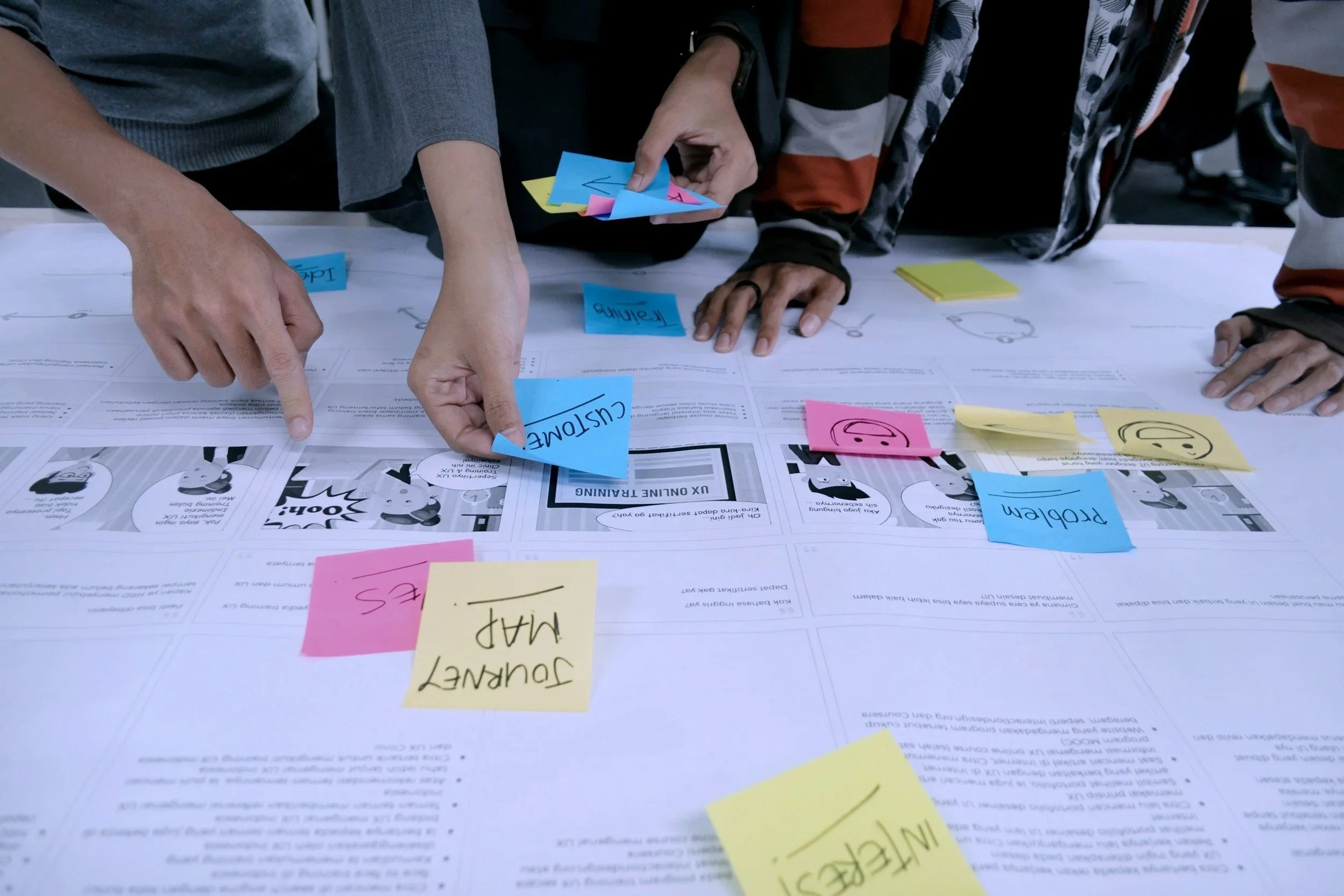 People working on a table with colorful sticky notes and printed documents, pointing and discussing.