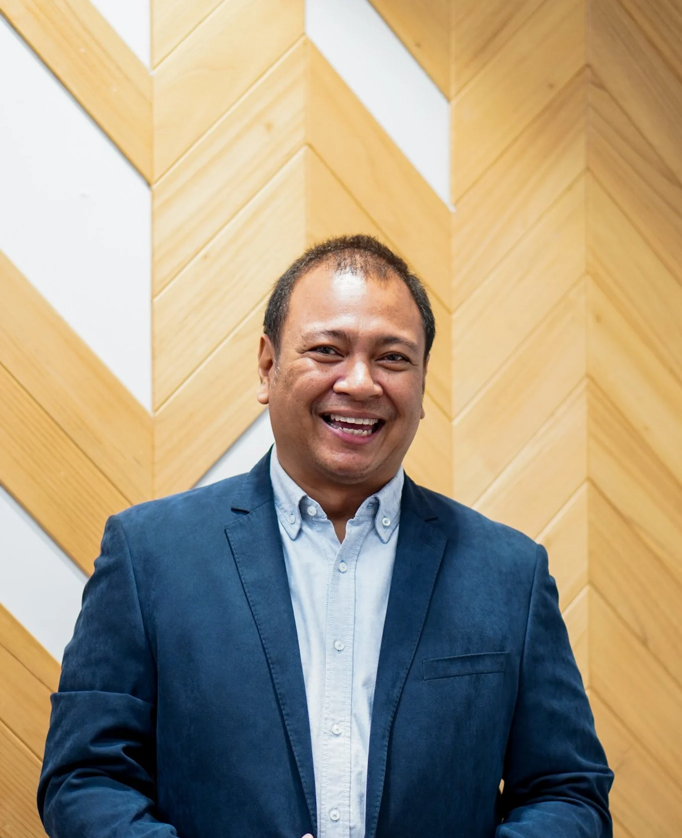 A smiling Balinese man in a blue blazer and light blue button-up shirt standing in front of a wooden wall with geometric pattern.