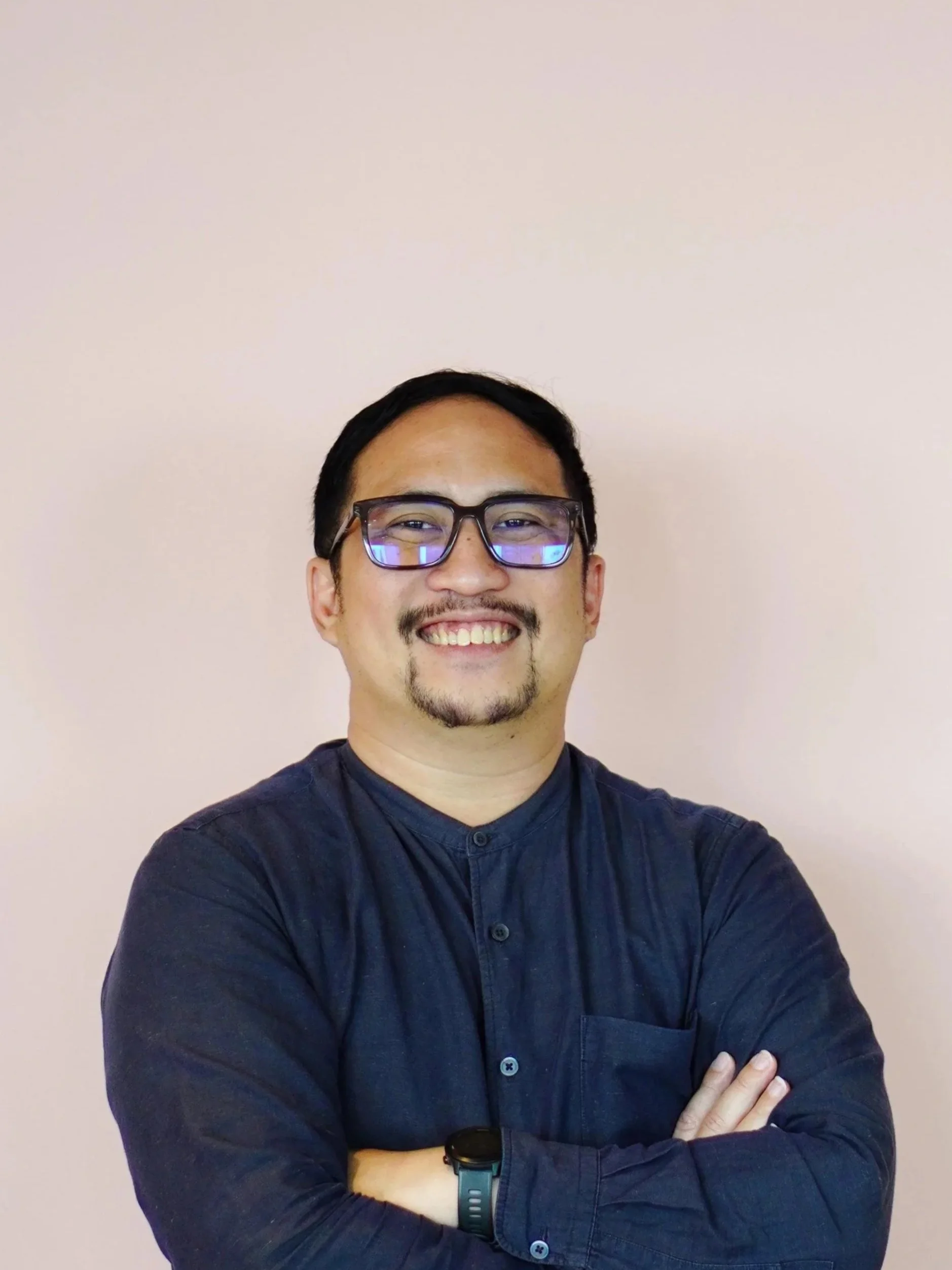 A Indonesian man wearing glasses and a dark blue button-up shirt smiling with his arms crossed, standing in front of a light pink wall.