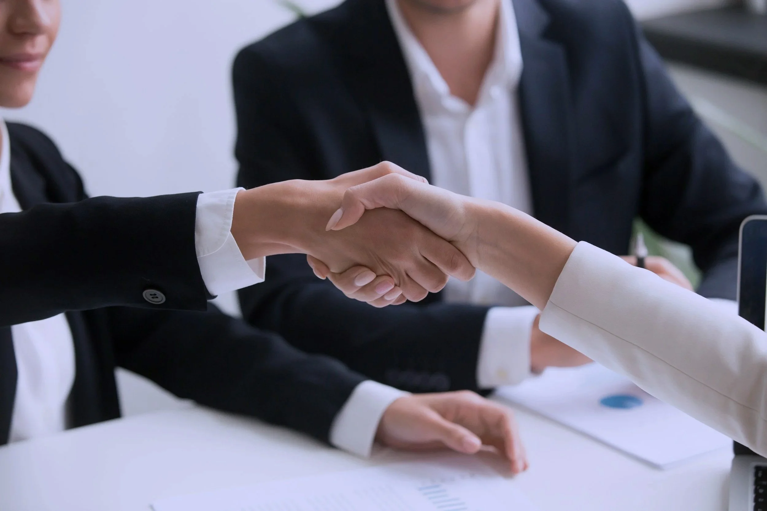 Two women in business attire shaking hands during a meeting in an office.