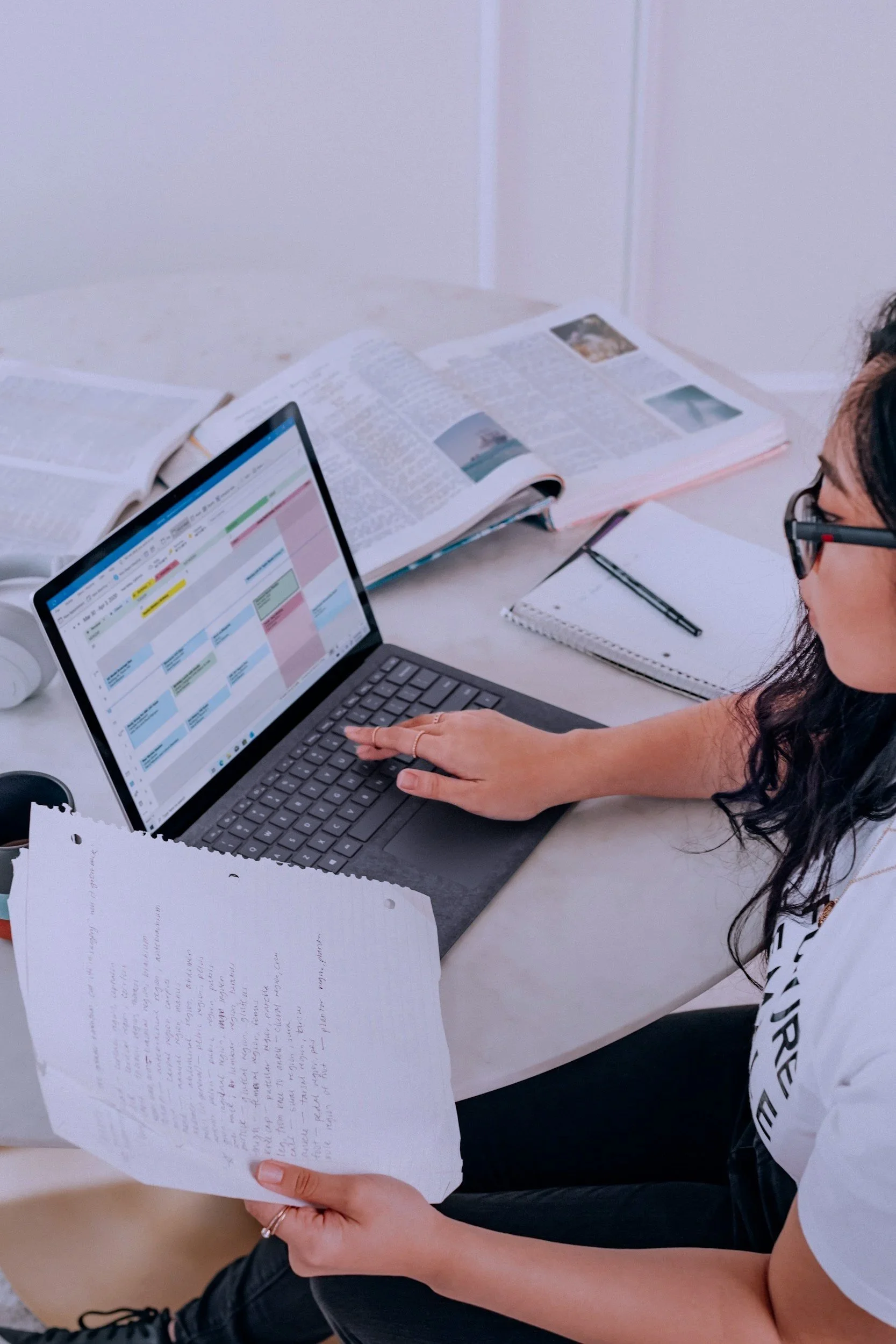 A person holding handwritten notes and working on a laptop at a desk with open textbooks, a notebook, and a pen.