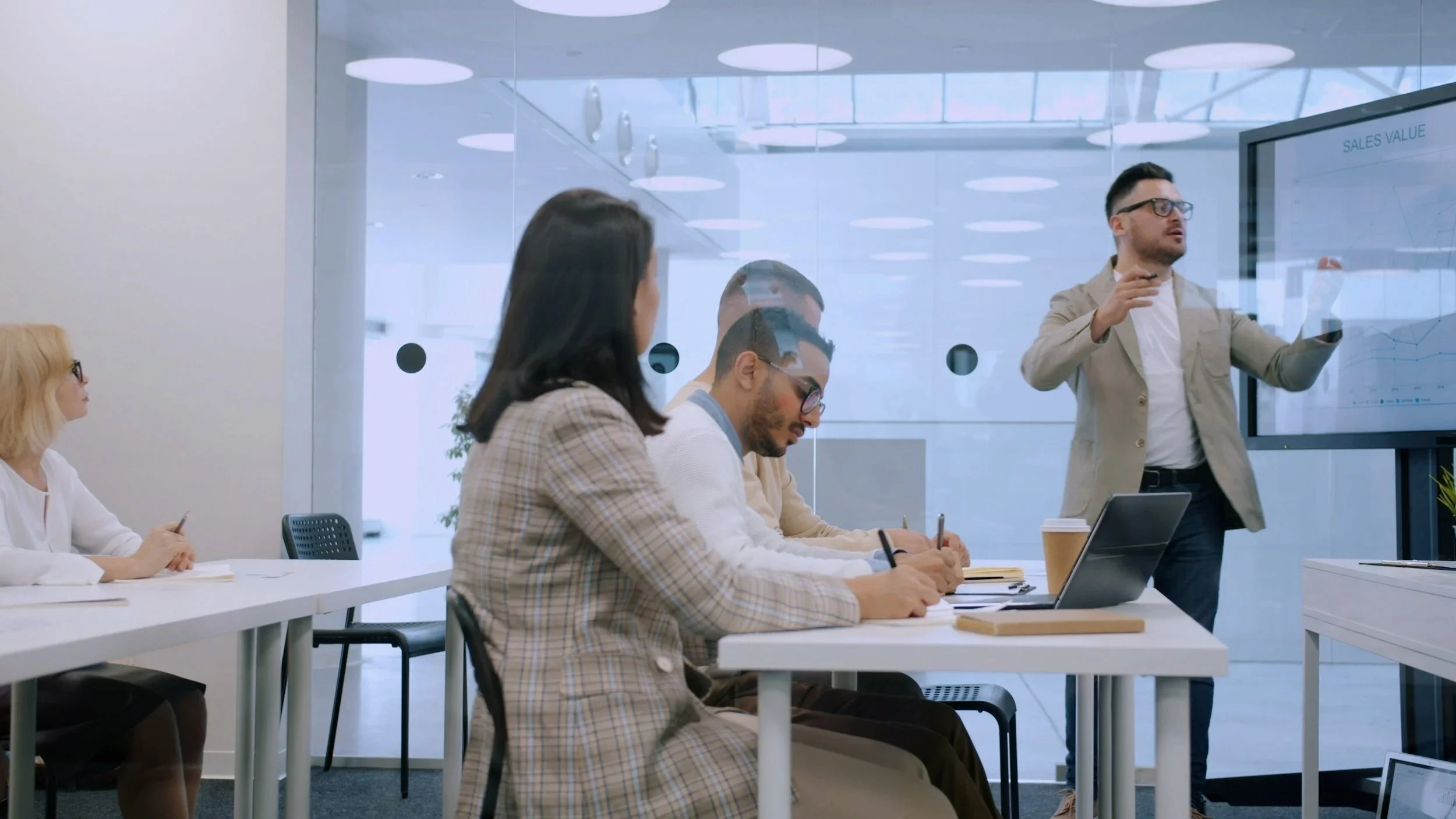 People are sitting at a wooden table in a meeting room, engaging in a discussion. One person is gesturing with both hands, and laptops, notebooks, and a smartphone are on the table.