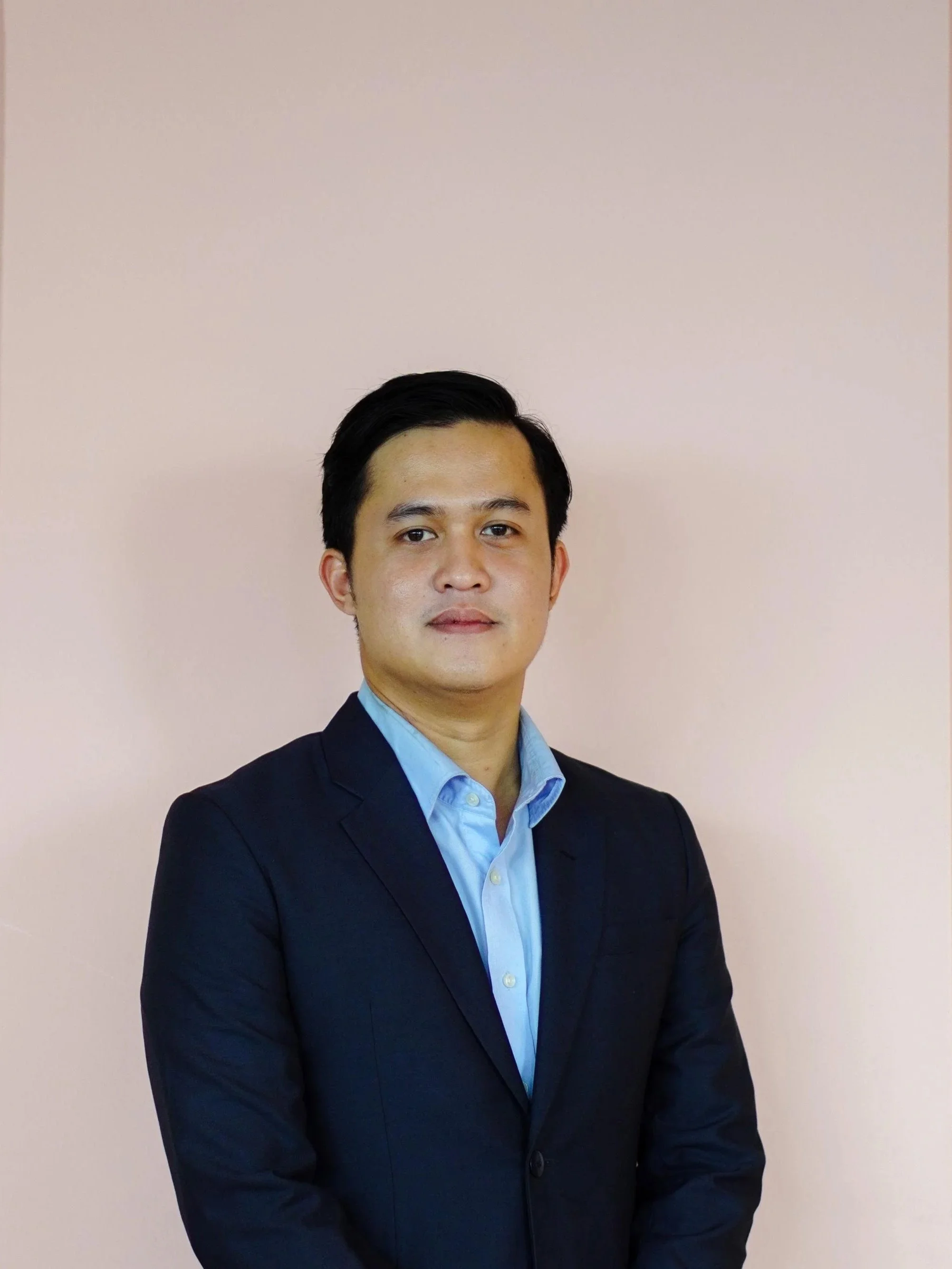 An Indonesian man wearing a navy jacket and blue shirt standing in front of a pink wall.