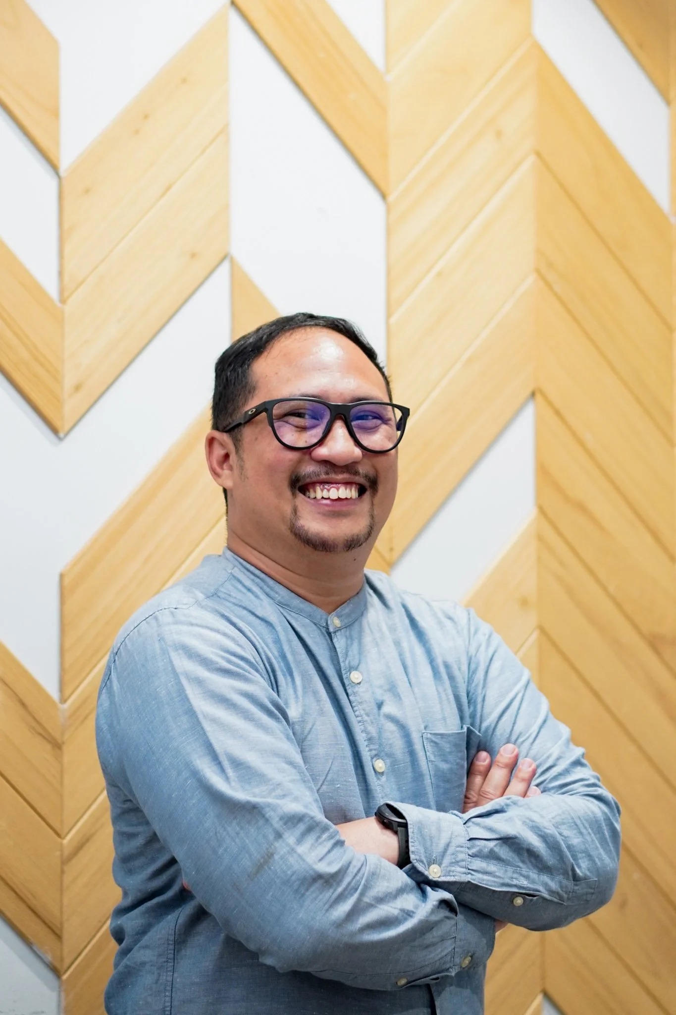 A Indonesian man wearing glasses and a light blue button-up shirt smiling with his arms crossed, standing in front of a wooden geometric wall pattern.