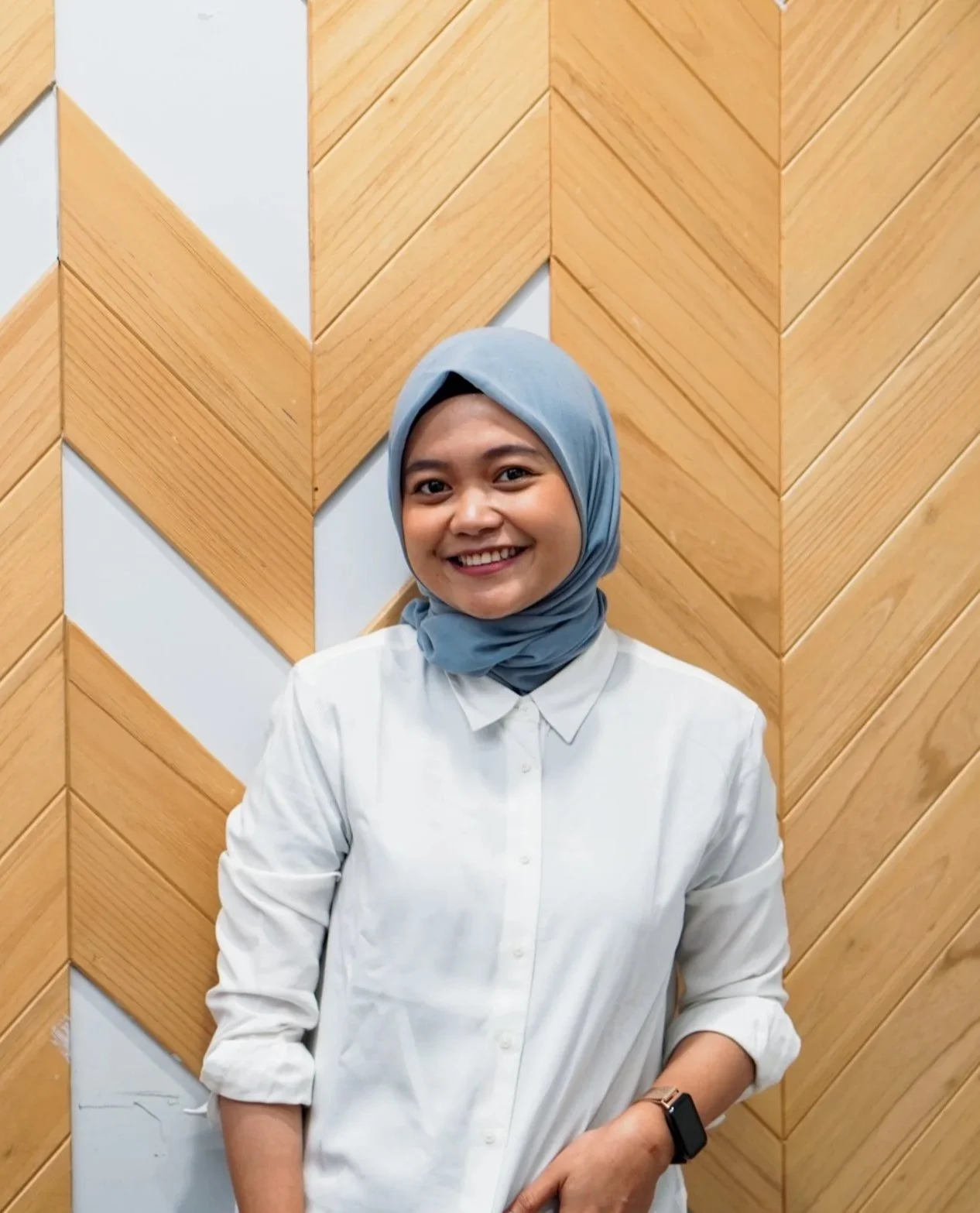An Indonesian professional woman smiling, wearing a light blue hijab and white button-up shirt, standing in front of a wooden wall with a chevron pattern.