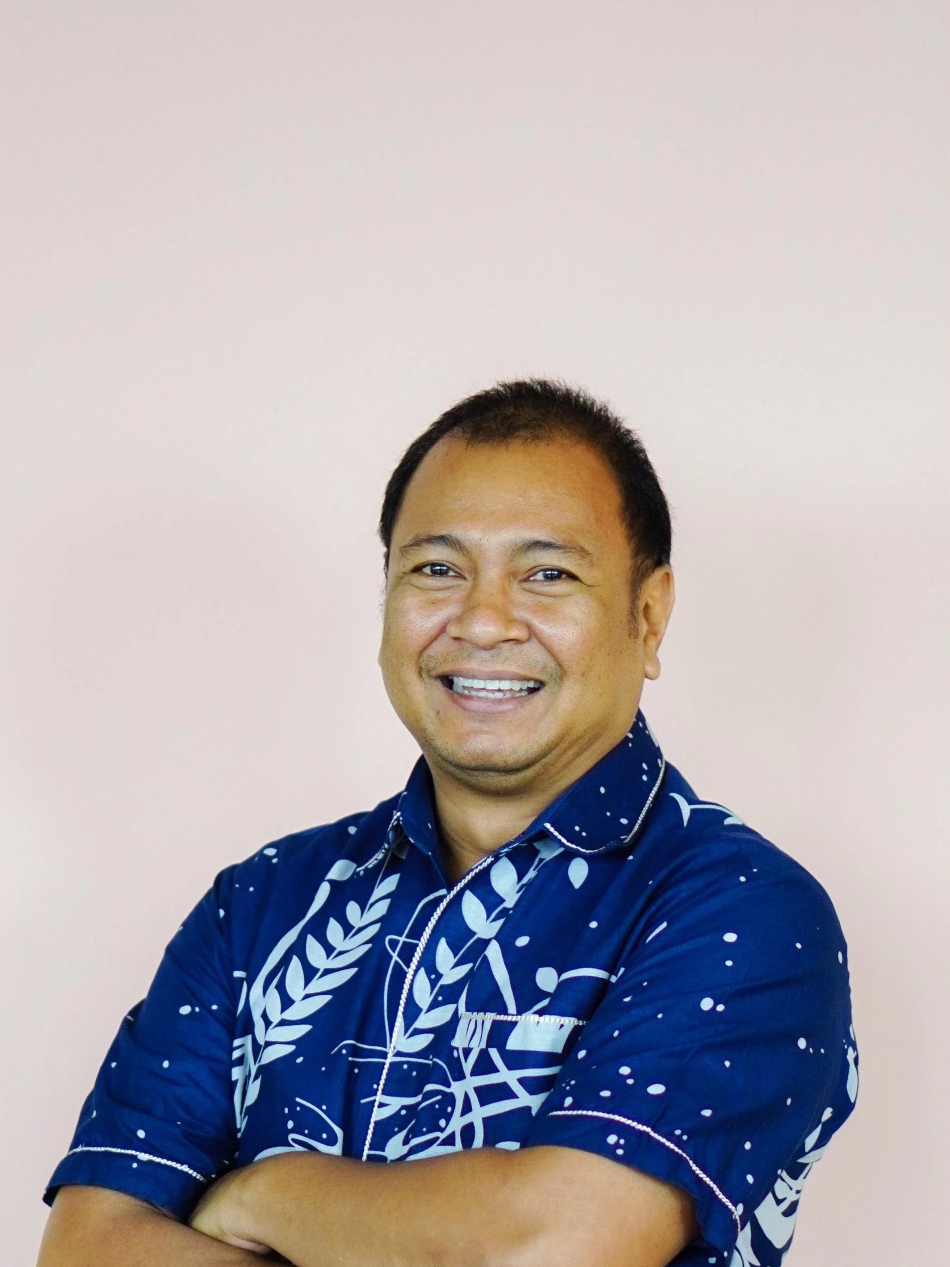 A smiling Balinese man in a blue batik shirt standing in front of a light pink wall.