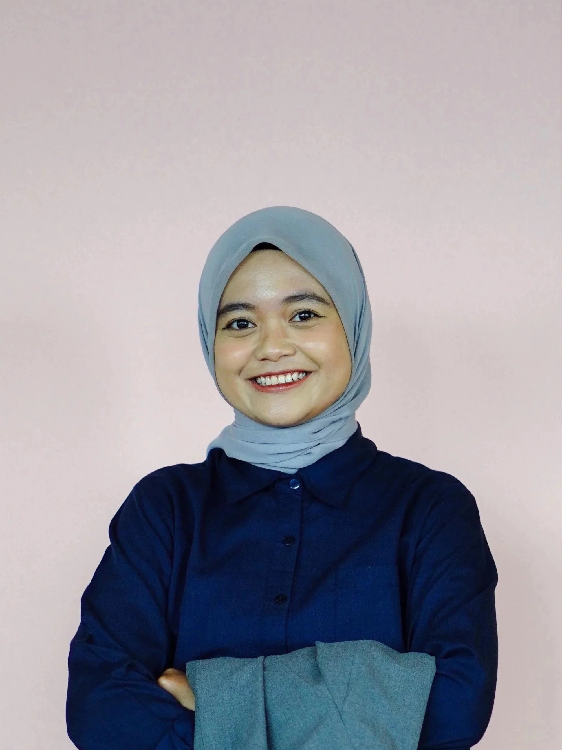 An Indonesian professional woman smiling, wearing a light blue hijab and white button-up shirt, standing in front of a wooden wall with a chevron pattern.