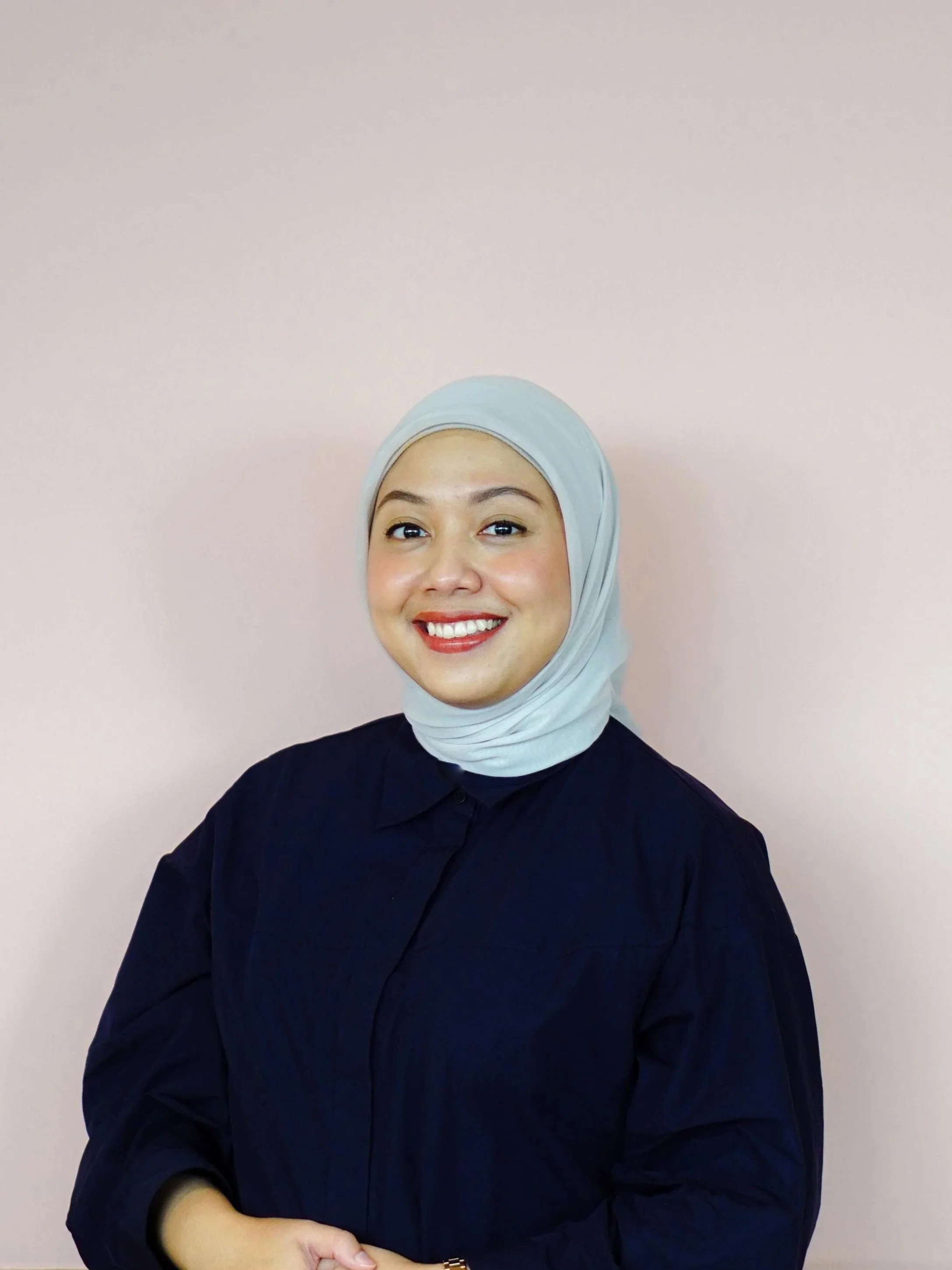 An Indonesian woman in a grey hijab and dark blue shirt smiling in front of a pink wall.