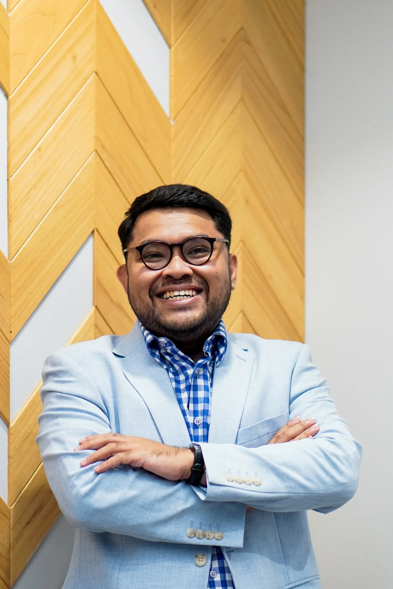 A Indonesian man in a light blue blazer and glasses smiling with arms crossed in front of a wooden wall.