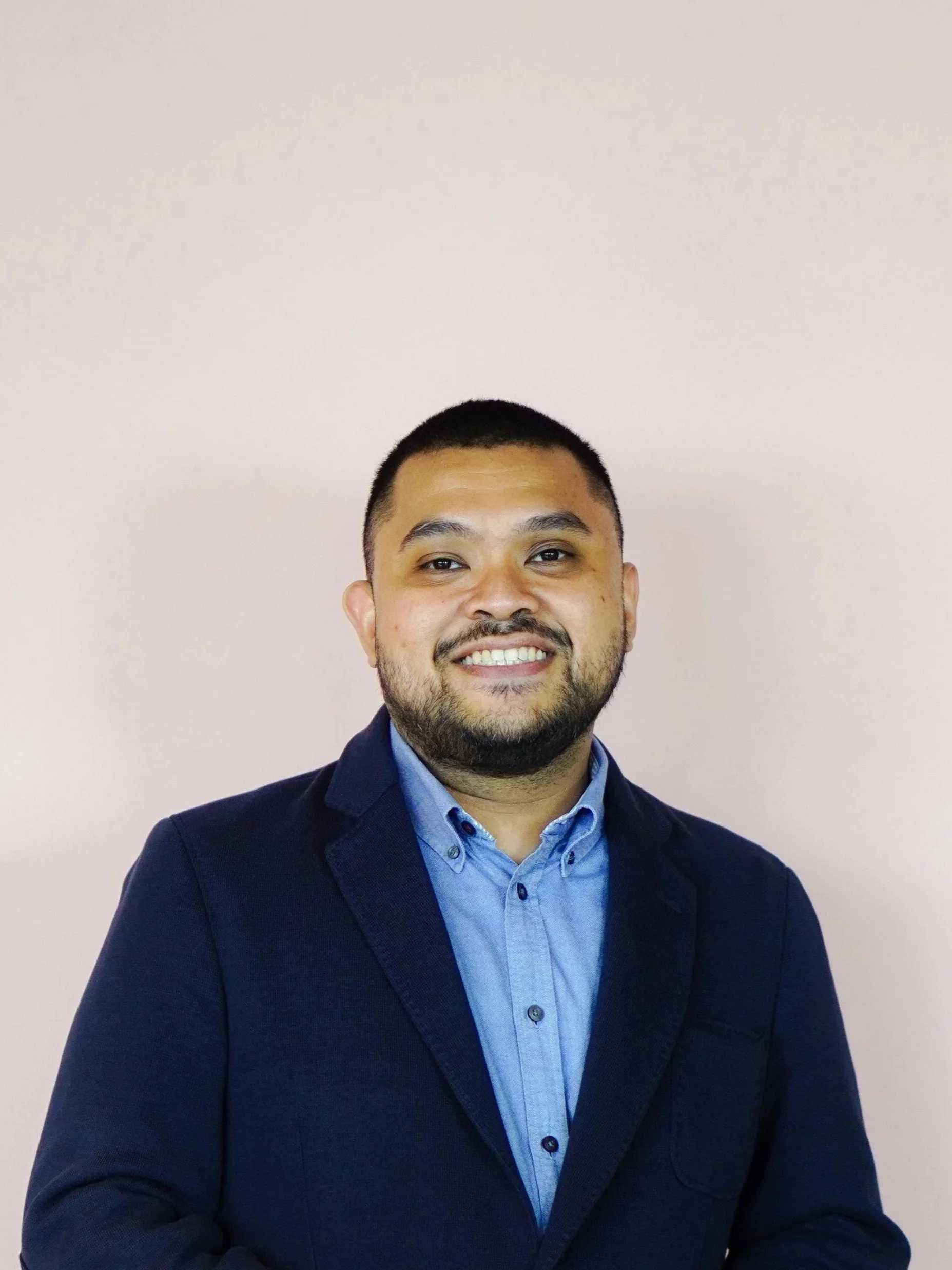 A Indonesian man in a dark blue blazer, blue shirt, and glasses smiling in front of a pink wall.