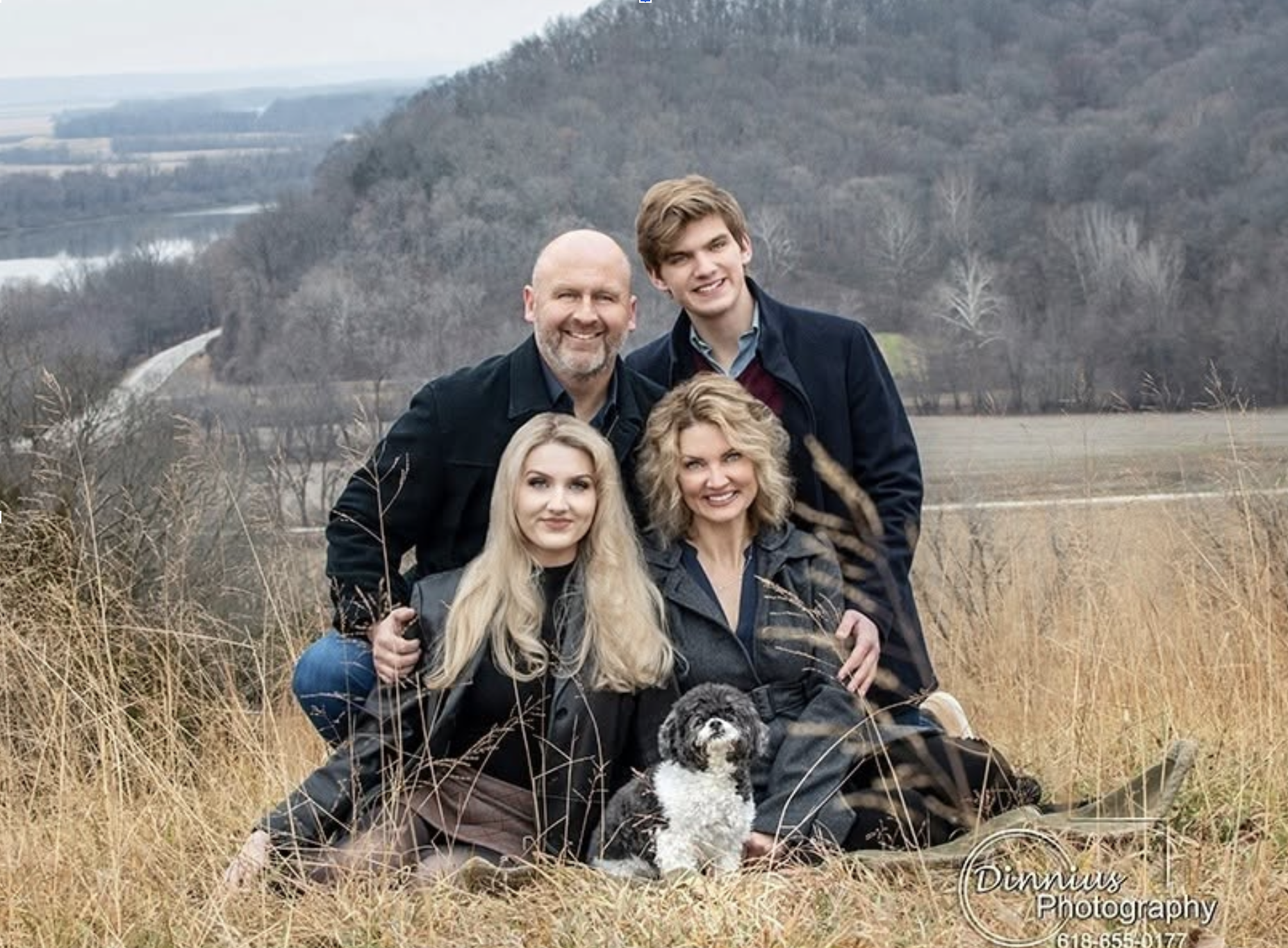 A group of five people and a dog posing outdoors in a field, with hills and a river in the background during fall or winter.