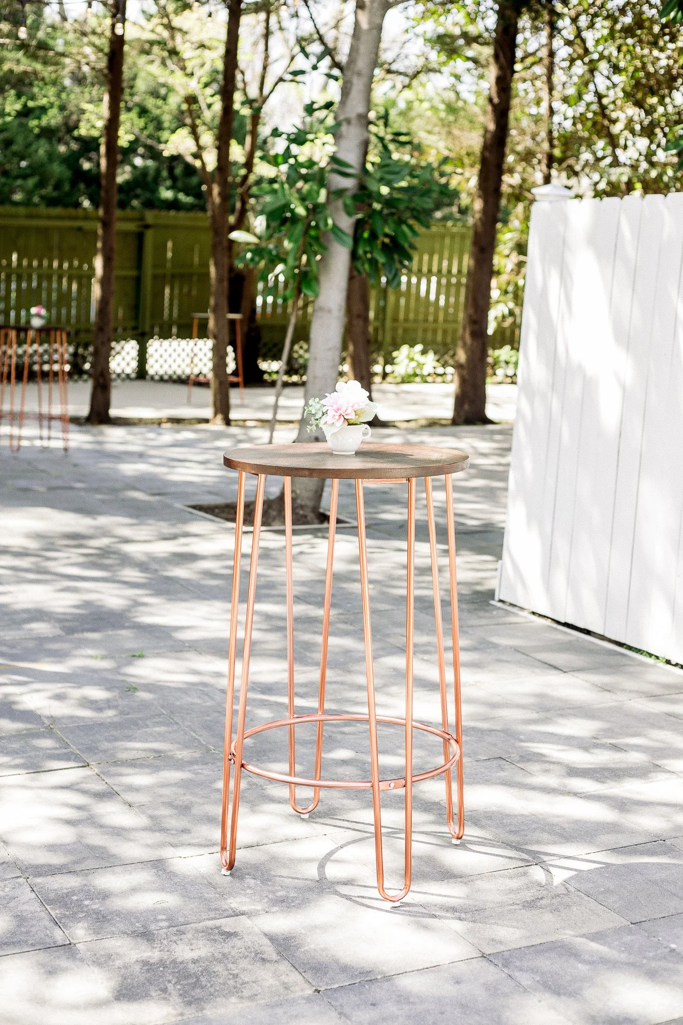 A round wooden table with copper legs holding a small white vase with pink and white flowers, set outdoors on a stone patio with trees and a white fence in the background. Garden Wedding venue Atlanta Georgia The Wildflower 301