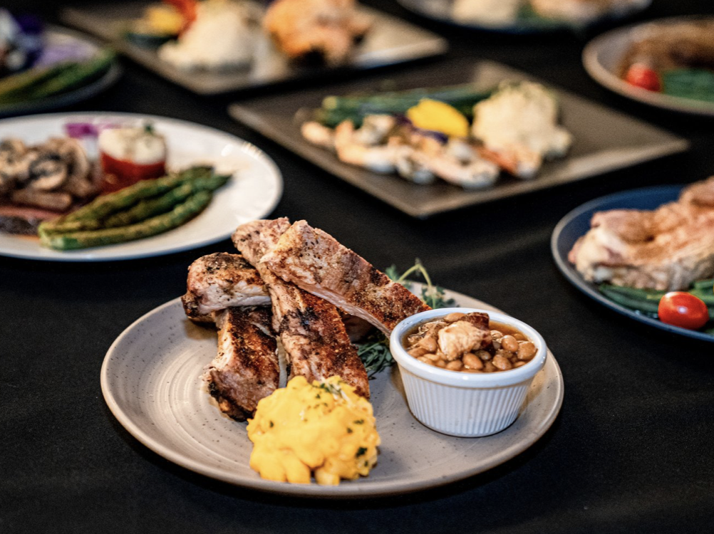 Plate with grilled pork ribs, mashed potatoes, and baked beans, with other dishes in the background on a black tablecloth. Historic Mansion Garden Wedding venue Atlanta Georgia The Wildflower 301