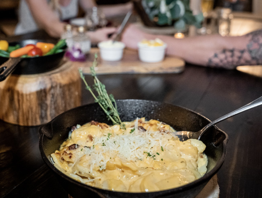 A close-up of a skillet of cheesy pasta garnished with thyme on a dark wooden table, with blurred vegetables and people in the background. Historic Mansion Garden Wedding venue Atlanta Georgia The Wildflower 301