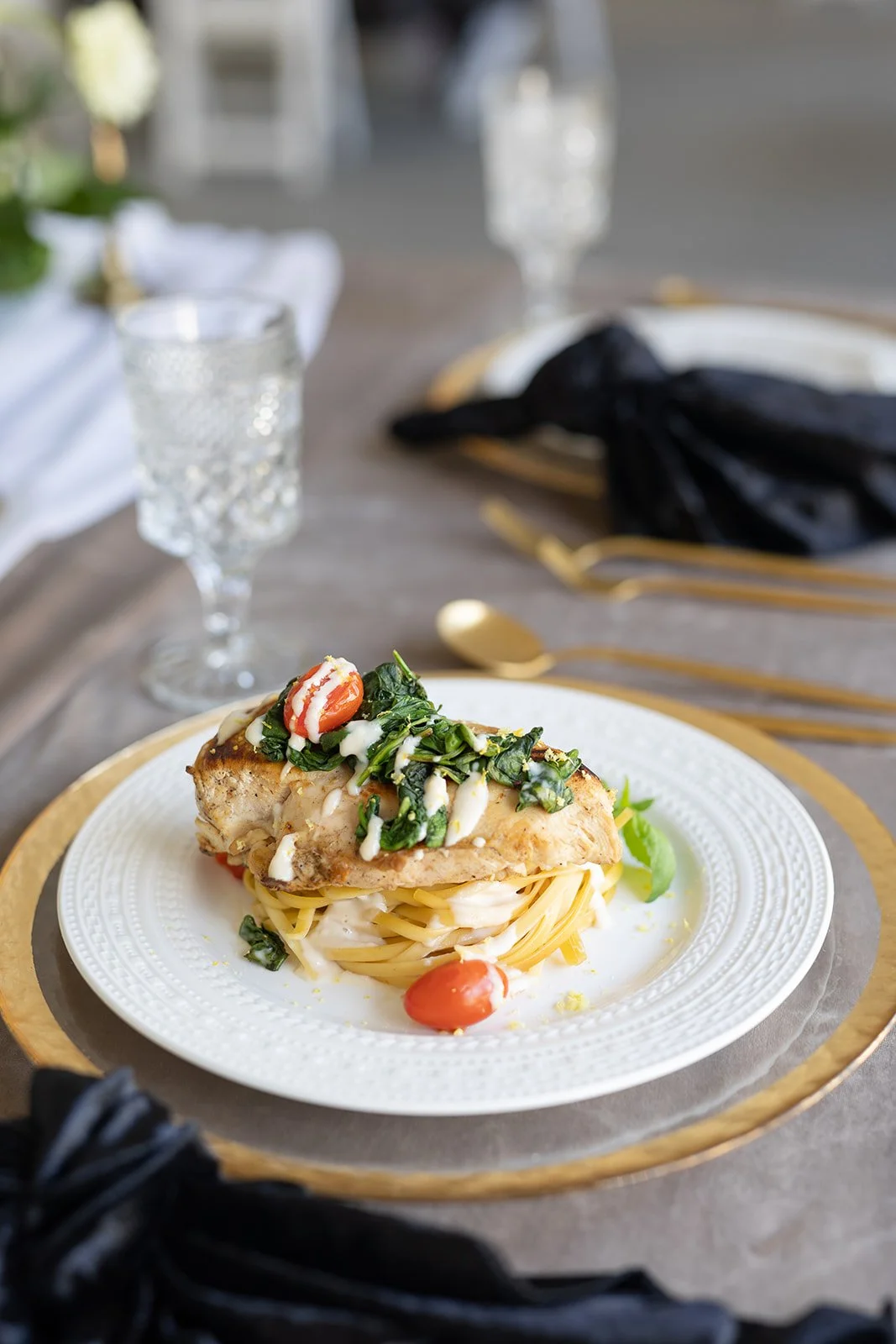 A plated serving of pasta topped with cooked greens, cherry tomatoes, and a piece of seasoned chicken, on a white plate with gold accents, set on a table with gold and black cutlery and crystal glasses. Garden Wedding venue Atlanta Georgia The Wildfl