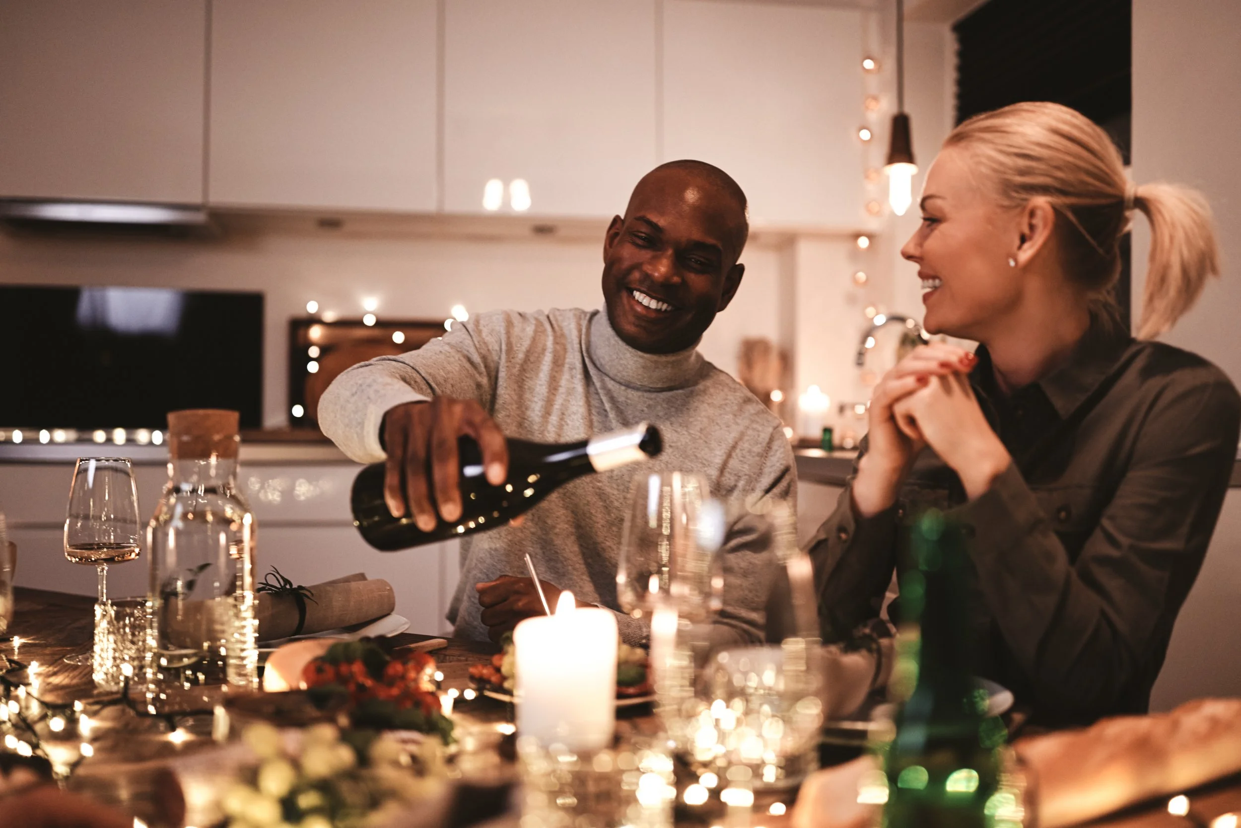 A man pouring wine for a woman during a dinner party in a warmly lit kitchen adorned with string lights, with food, drinks, and candles on the table. Garden Wedding venue Atlanta Georgia The Wildflower 301
