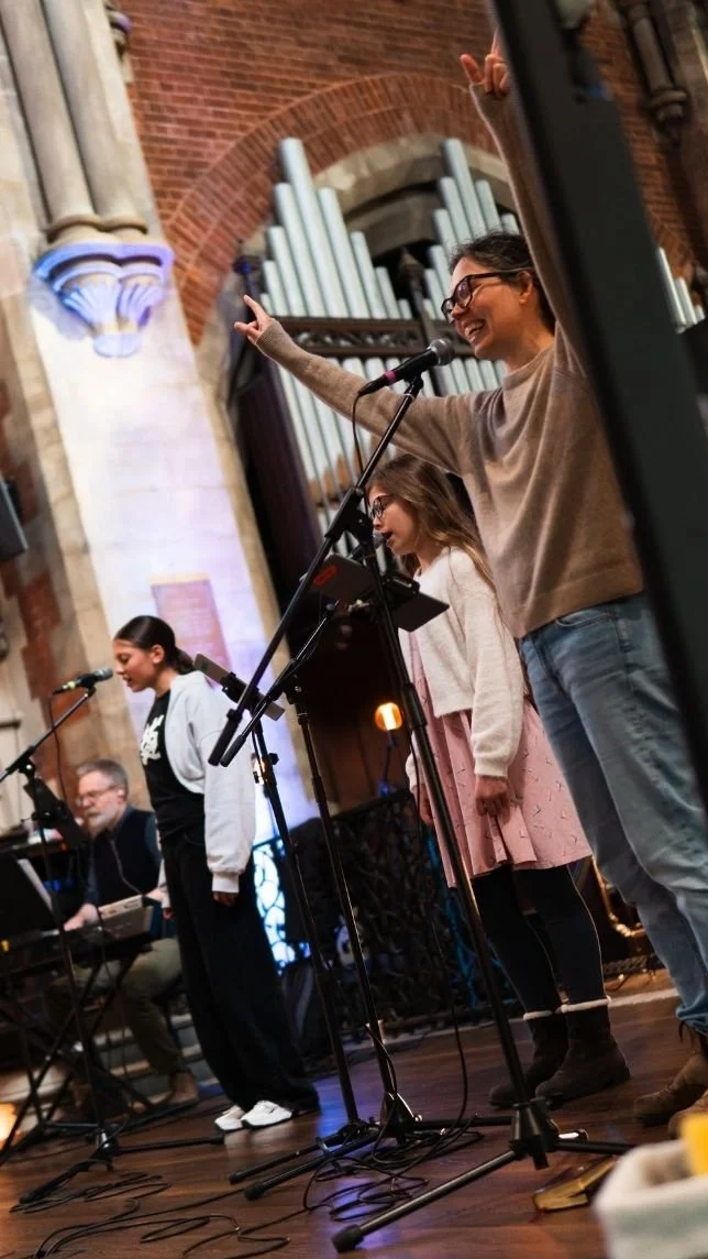 Musicians perform on a church stage with microphones and organ pipes in the background.