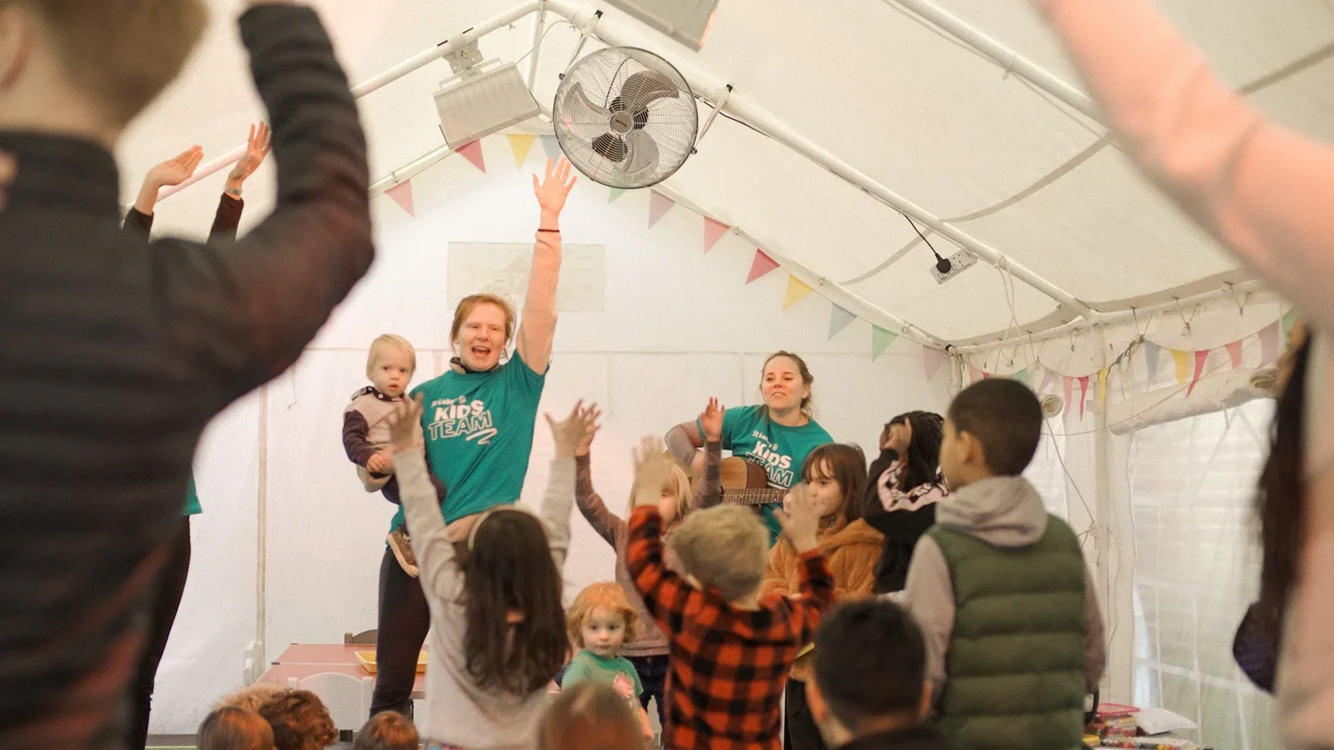 Children and adults with raised hands singing and dancing during a kids' group activity inside a decorated tent.