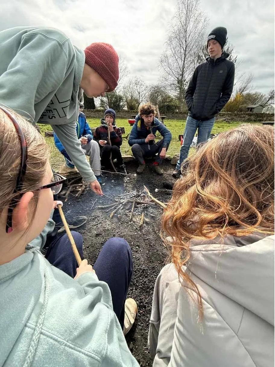 Children gathered outdoors around a small campfire pit, tending sticks and roasting food in a grassy yard.