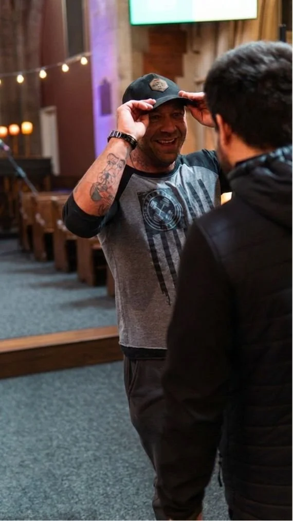 Smiling man adjusting a black baseball cap while talking to another person in church
