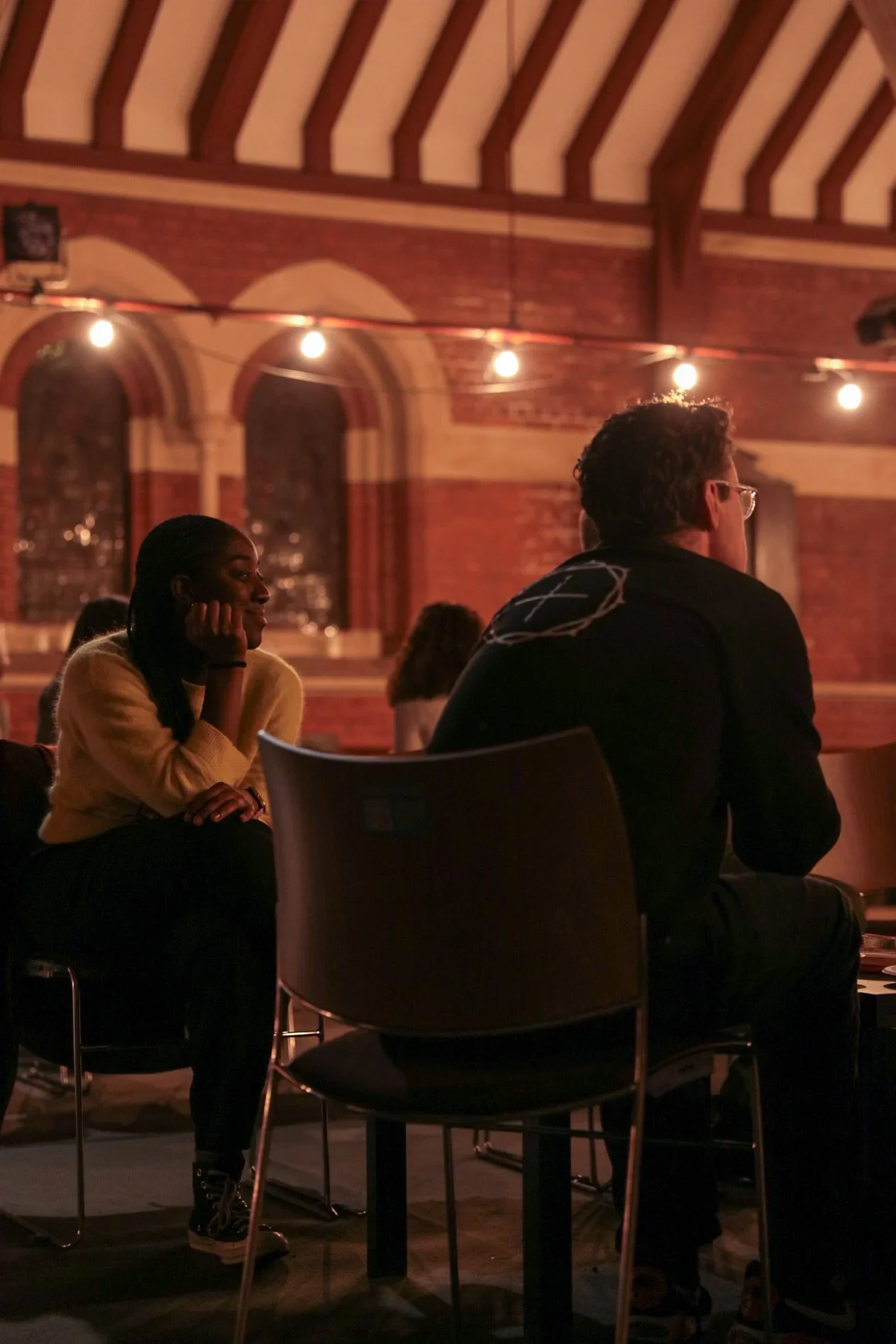 People socializing in a warmly lit historic brick venue with arched windows and string lights